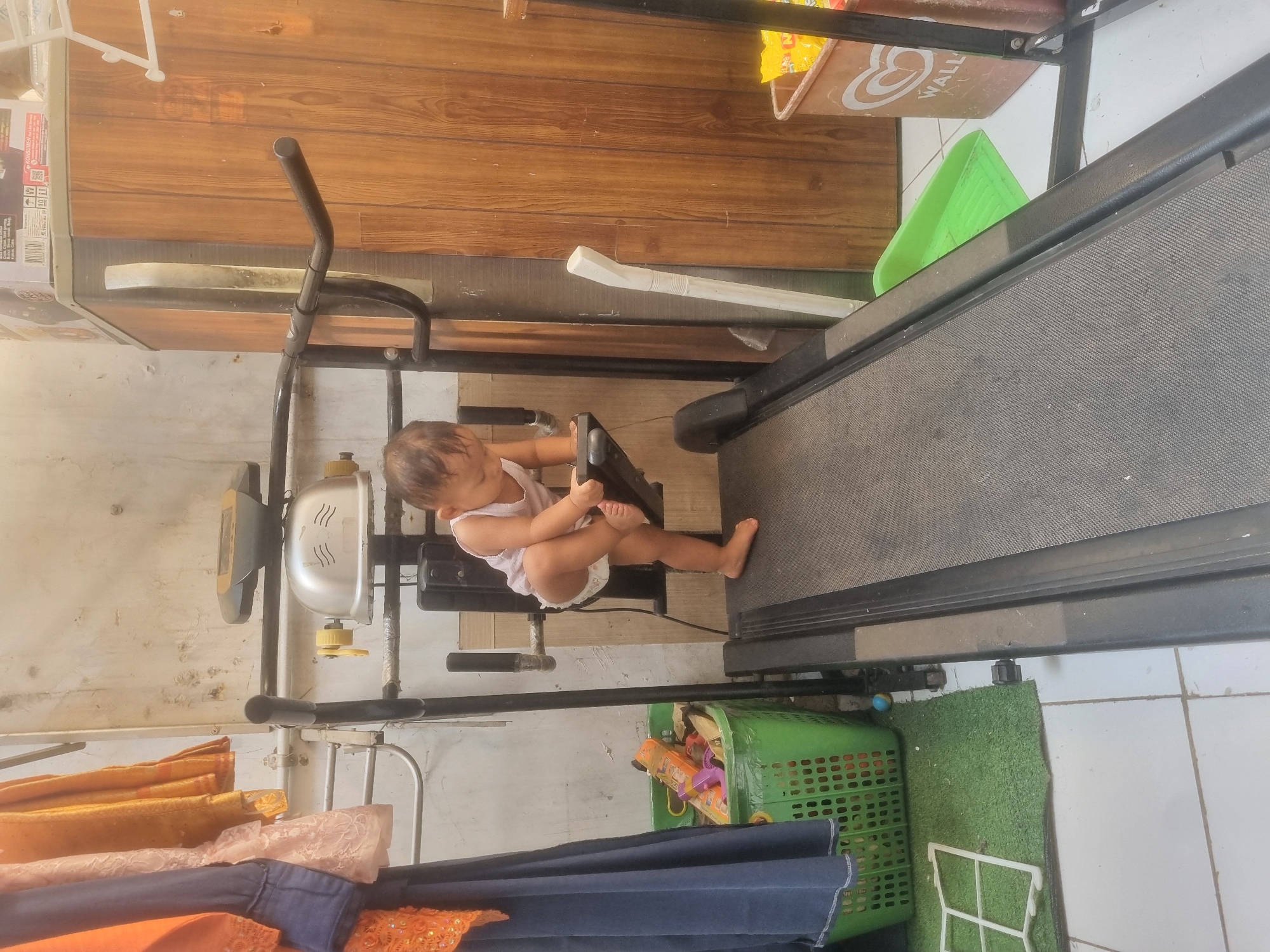A toddler, wearing a white tank top and a diaper, stands on a black treadmill, holding onto the handlebars. The toddler's left leg is bent, with the foot resting on the treadmill belt, and the right leg is extended. The treadmill is in an outdoor area, with a white tiled floor and a textured green mat to the left. A green plastic basket filled with toys is also visible on the mat. In the background, there is a wooden wall, some hanging clothes, and a white plastic object leaning against the wall. The scene appears to be in a residential area in Probolinggo, Indonesia.
