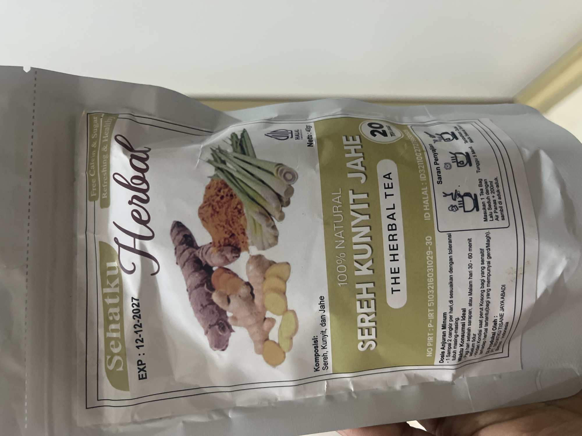 A white, resealable pouch of "Sehatku Herbal" tea is held by a person's hand in the lower left of the frame. The pouch displays Indonesian text and imagery of turmeric, ginger, and lemongrass. The expiration date is visible as "EXP : 12-12-2027." The pouch is oriented vertically, with the top slightly folded over. The background is a plain, light-colored wall. There are no discernible elements in the image that would indicate its location in Denpasar, Indonesia, or any specific city.