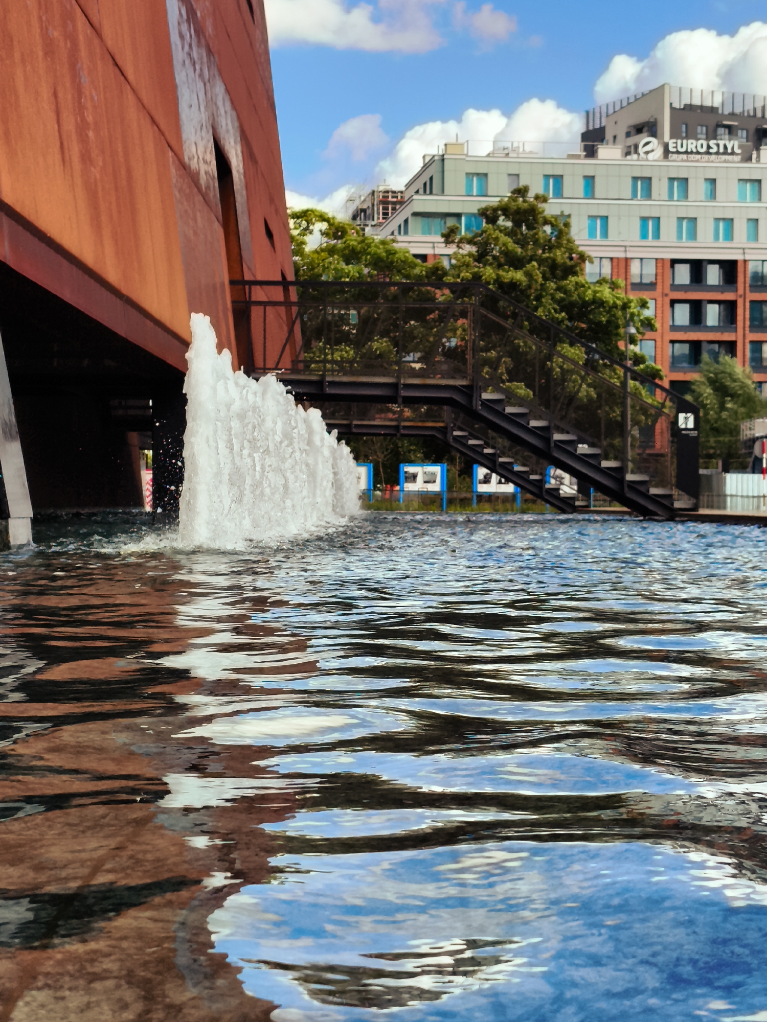 The image captures a contemporary architectural scene near the Motława River in Gdańsk, Poland. In the foreground, the rippling surface of a shallow pool reflects the sky, clouds, and surrounding structures, creating abstract patterns of light and color. A modern fountain in the mid-ground shoots a vigorous spray of water upwards, adding a dynamic element to the scene.
To the left, a section of a large, rust-colored building dominates the frame, its angled facade and industrial aesthetic suggesting a modern design. Beyond the fountain, a black metal staircase with open treads leads upwards, providing access to a higher level. The staircase is partially obscured by lush green trees, which also screen a row of modern apartment buildings in the background.
These background buildings are characterized by their multi-story design, clean lines, and a mix of materials including brick and light-colored facades. One prominent building displays the "EURO STYL GRUPA DEWELOPMENT" logo, indicating a commercial development. The sky above is a bright blue, dotted with fluffy white clouds, suggesting a pleasant, possibly sunny day. The overall impression is one of urban renewal and modern architecture integrated with water features and greenery.