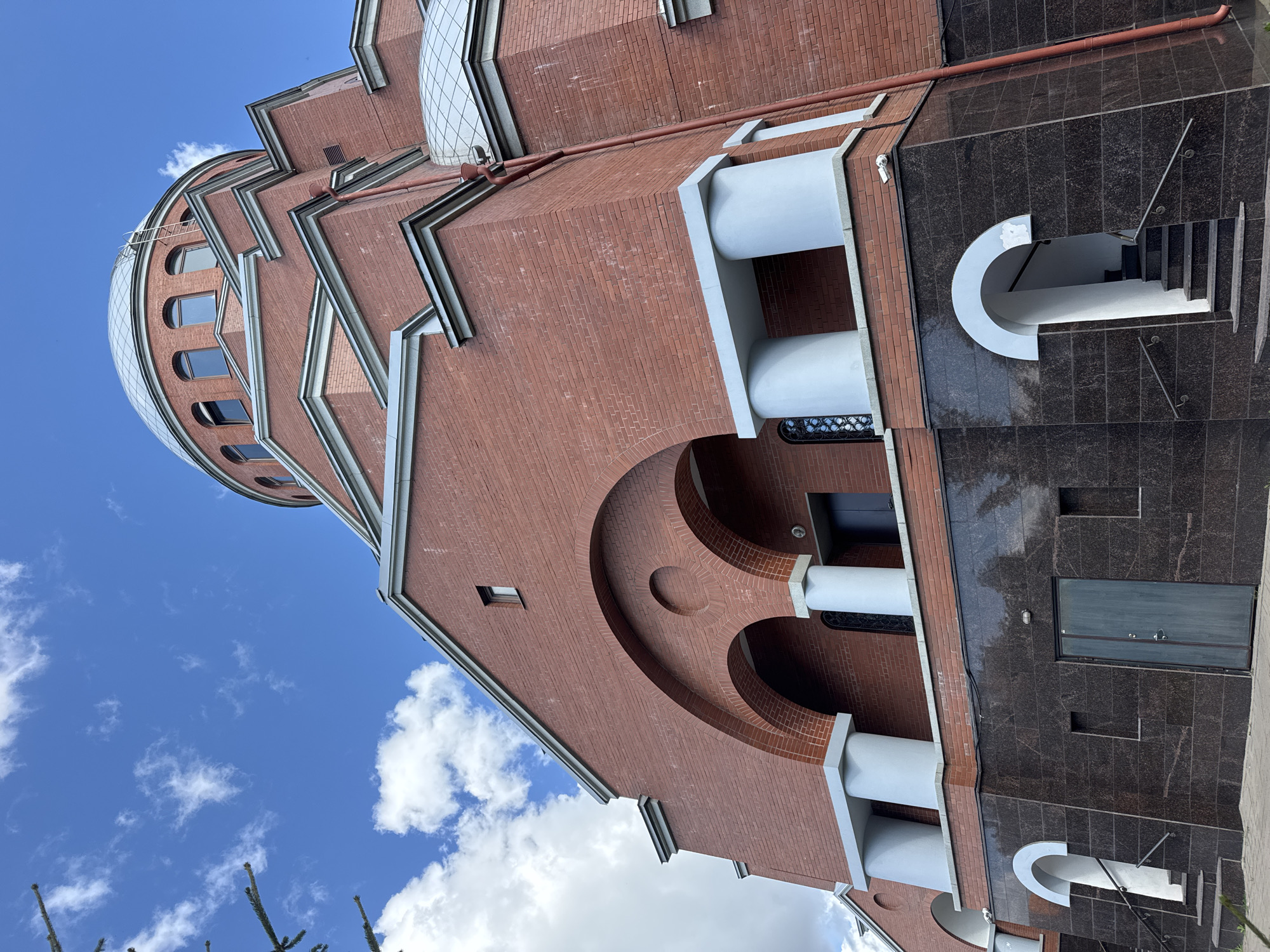 The image shows a large, modern, red brick building with a silver dome, likely a church or religious institution, located in Sinyavino, Russia. The building features arched entrances, white columns, and dark granite detailing at the base. A red downspout runs vertically along the side of the building. The sky is blue with scattered white clouds. There are no people visible in the image. The perspective is from a low angle, looking up at the structure.