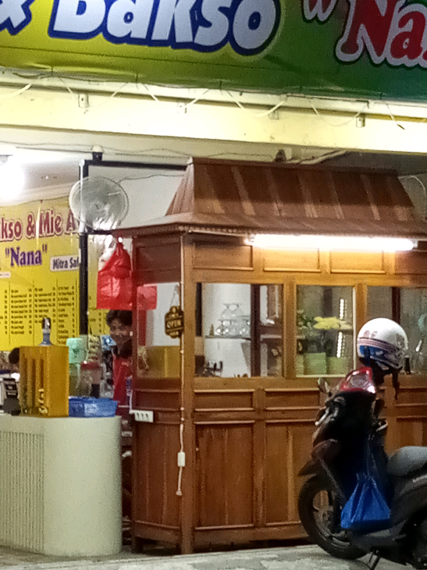 A street-level view shows a food stall at night. The stall has a wooden counter and a wooden roof with a fluorescent light. A motorcycle is parked in front of the stall, with a blue bag hanging from its handlebars. A helmet rests on the back of the motorcycle seat. A person wearing a red shirt is visible behind the counter, partially obscured by a red plastic bag hanging from a hook. A sign above the stall reads "Bakso & Mie Ayam 'Nana'" and "Mitra Sa...". The stall is located in the city of Bekasi, Indonesia. The overall lighting suggests it is nighttime, and the scene is typical of a street-side food vendor.