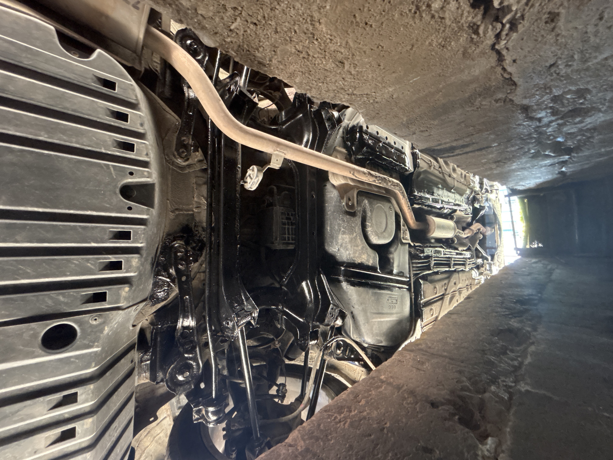 The image shows the underside of a vehicle, possibly a car, viewed from below. The vehicle is positioned over a pit, as evidenced by the concrete ledge visible on the right side of the frame and the bright light shining from the far end of the pit.
The vehicle's undercarriage is visible, including exhaust pipes, suspension components, fuel tank, and a protective plastic underbody panel on the left. The components appear to be coated with a thick, black substance, likely rust inhibitor or undercoating.
The image perspective is looking upwards, providing a clear view of the vehicle's undercarriage and the concrete ceiling above.
The location of Zlatograd, Bulgaria cannot be confirmed, and there are no people visible in the image.