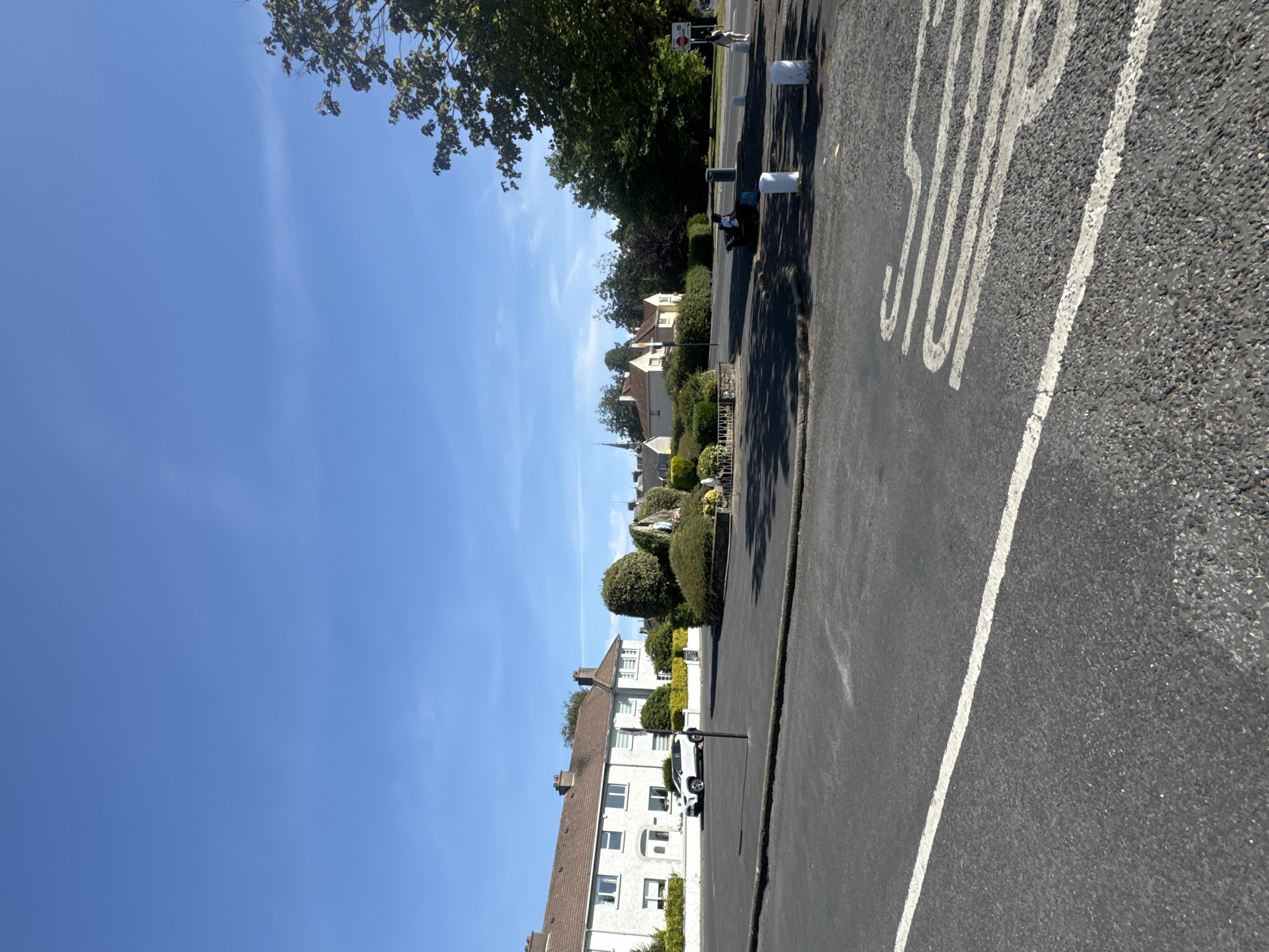 The image depicts a street scene under a clear blue sky in Waterford, Ireland. In the foreground, a dark asphalt road with a white line marking is visible. Further back, a small roundabout features a sculpted figure, possibly a religious statue, surrounded by manicured bushes. To the left, a white car is parked in front of a row of terraced houses with red-tiled roofs and white facades. The houses appear to be two stories high with multiple windows. On the right side of the roundabout, a bench is occupied by two individuals, one of whom is looking away from the camera. A trash can is situated near the bench, and several white bollards are spaced along the edge of the road. In the background, trees and more houses are visible, with a church spire rising above the rooftops. A traffic sign with a red circle and a white horizontal line is also present on the right. The overall scene suggests a quiet, residential area on a sunny day.