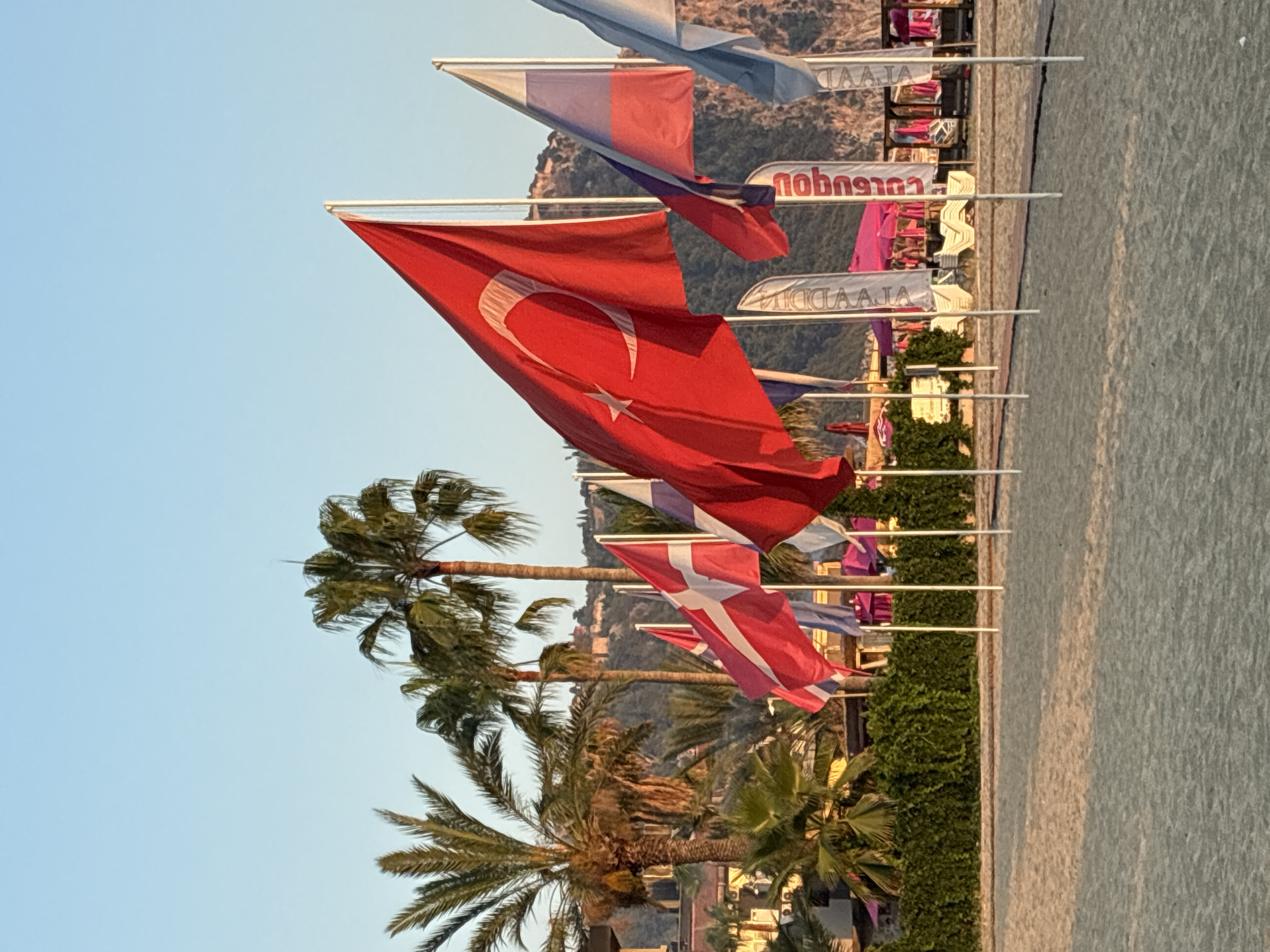 The image depicts a beach scene in Alanya, Turkey, under a clear blue sky. Several national flags are prominently displayed on poles along the beach, including the Turkish flag, a Danish flag, and a Russian flag. Palm trees are visible in the background, alongside buildings and a hillside. Beach chairs and umbrellas are set up on the sand, suggesting a resort area. The sun is low in the sky, casting a warm light on the scene. No people are clearly visible in the frame, and no specific actions or interactions are depicted. The environment is predominantly sandy beach, vegetation, and clear sky, with distant hills.