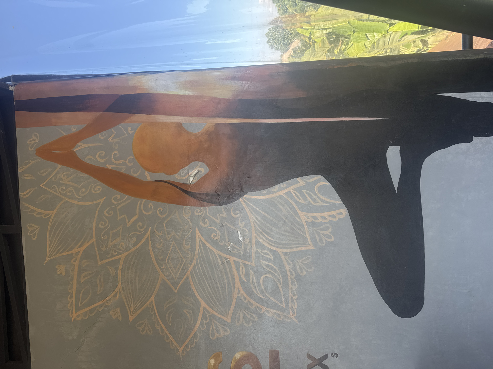 A mural on a grey wall depicts a silhouette of a person in a yoga pose, with their arms raised above their head and hands clasped. Behind the silhouette, a large, light orange mandala design is painted. To the right of the silhouette, vertical stripes of pink, brown, and black are painted. The mural is located in the city of Ubud, Indonesia, and the mural's style and imagery, such as the mandala and yoga pose, are consistent with the cultural and spiritual themes often found in Ubud. The mural is visible next to a window, through which lush green foliage and a clear blue sky can be seen. Part of a black metal frame is visible in the foreground on the right side of the image.