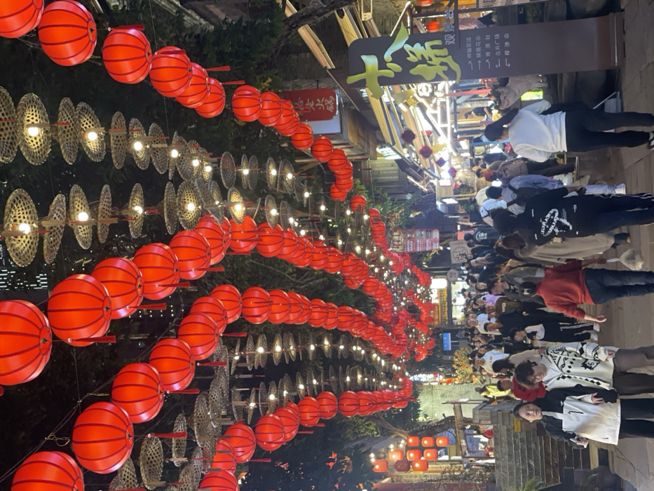 This image depicts a densely populated pedestrian street illuminated by numerous red lanterns and decorative lights, located in Yuzhong District, China. Overhead, a canopy of bright red cylindrical lanterns, each with a tassel, is strung in multiple parallel rows across the thoroughfare. Interspersed between these lanterns are strings of smaller lights housed within woven, saucer-shaped structures that resemble straw hats.
Below this illuminated canopy, a large crowd of individuals, primarily adults, walks along the street. They are dressed in casual attire, including jackets, sweaters, and trousers. People are moving in various directions, though generally forward into the frame, indicating a busy thoroughfare. No specific interactions beyond general pedestrian movement are discernible.
Buildings with visible storefronts line both sides of the street. On the right, a prominent brown and gold sign with large Chinese characters and directional arrows is visible, indicating local points of interest. Further along on the right, a red sign with white Chinese characters, possibly identifying a "Hotpot" establishment, is also present. Trees with visible foliage are integrated into the streetscape, particularly on the left side and possibly intertwined with the overhead decorations. Steps leading upwards from the street are visible on the left side. The scene is illuminated by artificial light, suggesting evening or night conditions.