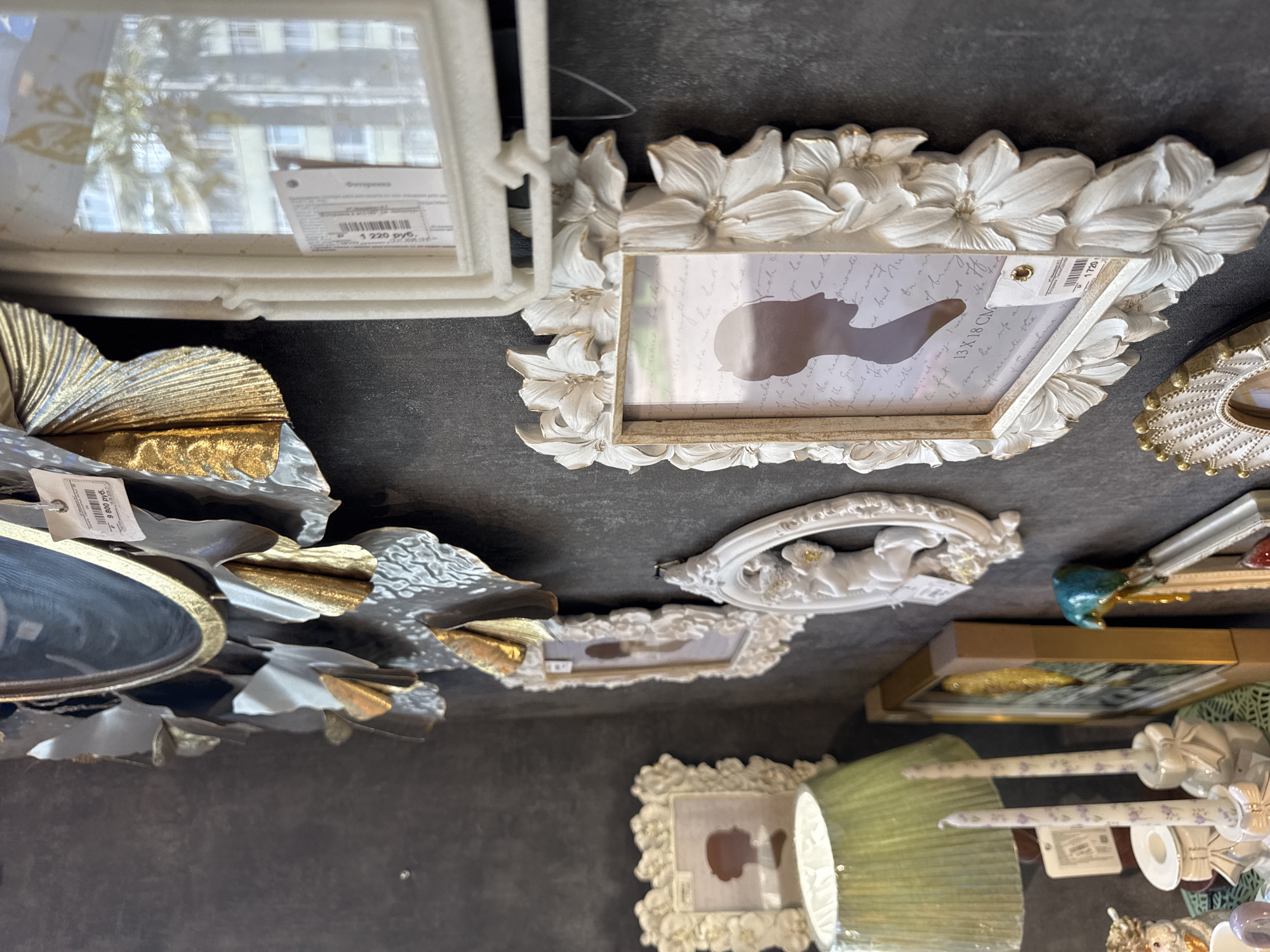 The image depicts a wall decorated with ornate picture frames and decorative items, likely in a shop or home interior. The overall style is vintage and elaborate, with a focus on white, gold, and darker tones.
Dominating the upper left is a large, decorative round mirror with a golden edge and elaborate, metallic petal-like accents in gold and silver. A price tag, partially obscured, is attached to this piece.
To the right of the mirror, a tall, rectangular white picture frame with a textured surface and decorative floral elements is hung. A price tag is visible on this frame as well. The glass of this frame reflects an outdoor scene, showing a building and palm trees, suggesting an urban environment.
Below and to the left of this frame, a smaller, square white picture frame with a similar floral design contains a silhouette of a woman's profile against a background with text.
Further down and to the left, a green lampshade of a table lamp is visible, casting a soft light. Two white candles with small purple floral patterns and tied with ribbons are positioned in front of the lamp.
In the lower right quadrant, another ornate white picture frame, this one oval-shaped and adorned with cherubic figures and floral motifs, is displayed.
Below this, a larger rectangular picture frame with a golden border contains a silhouette of a woman's profile on a textured background with text. This frame also has a price tag. A small, teal-colored bird figurine is perched on the edge of another decorative item to the right.
At the very bottom, parts of other decorative items are visible, including a circular frame with a sunburst pattern and gold accents, and ceramic figurines.
The background is a dark, textured wall, possibly painted concrete or stucco. The lighting appears to be a mix of ambient shop lighting and the light from the lamp.
The specific location in Sochi, Russia, is not discernible from the visual information present in the image.