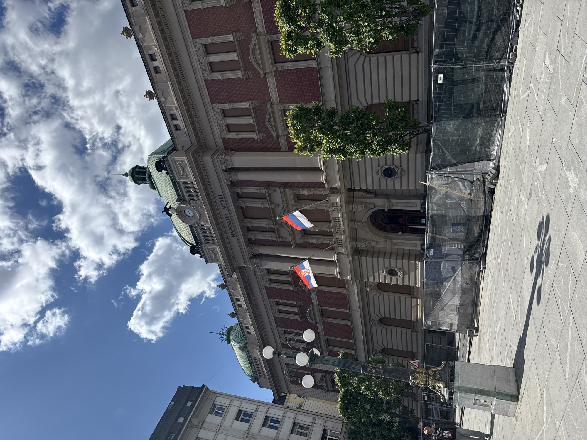 The image captures a street view in Belgrade, Serbia, focusing on the National Museum of Serbia building. Two flags, Russian and Serbian, are displayed on the building's facade. Construction fencing surrounds the base of the building, indicating ongoing work. A streetlamp stands in the foreground. The sky is visible, filled with clouds. A person is standing near the base of the streetlamp.