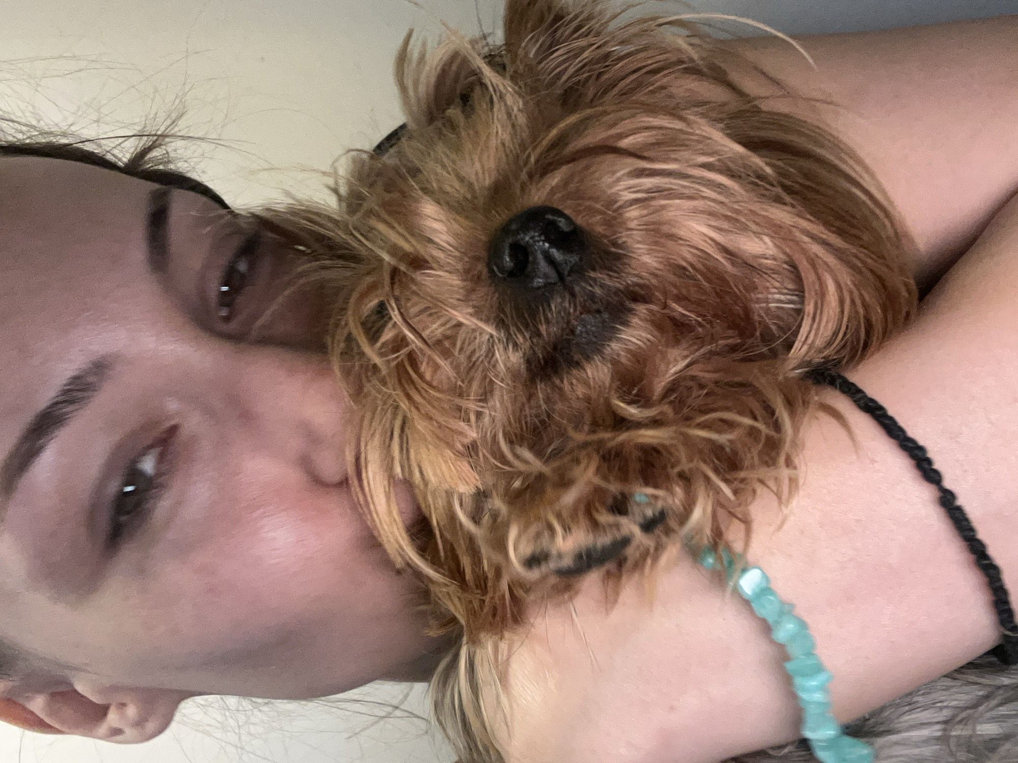 A selfie depicts a woman with brown eyes and hair lying on her back with a small, brown, scruffy dog resting on her chest and arm. The dog's black nose is prominent. The woman wears a black cord bracelet and a turquoise beaded bracelet on her left wrist. The background is blurred and indistinct, suggesting an indoor setting. There is no evidence in this image to suggest that the photo was taken in Burgas, Bulgaria.