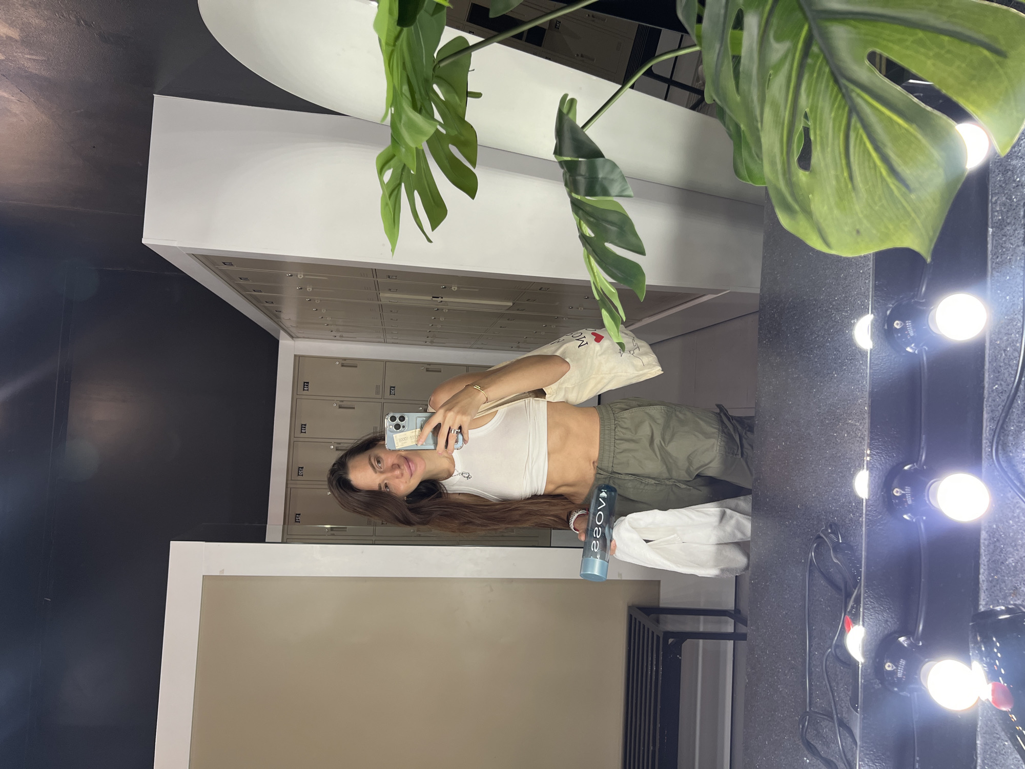 A woman is taking a selfie in front of a mirror in what appears to be a locker room. The locker room has a beige-painted wall, silver lockers, and a black-painted wall. The woman is holding a smartphone in her left hand and a water bottle in her right hand. She is wearing a white tank top and green pants. The mirror is surrounded by light bulbs. A green plant is situated on top of the mirror.