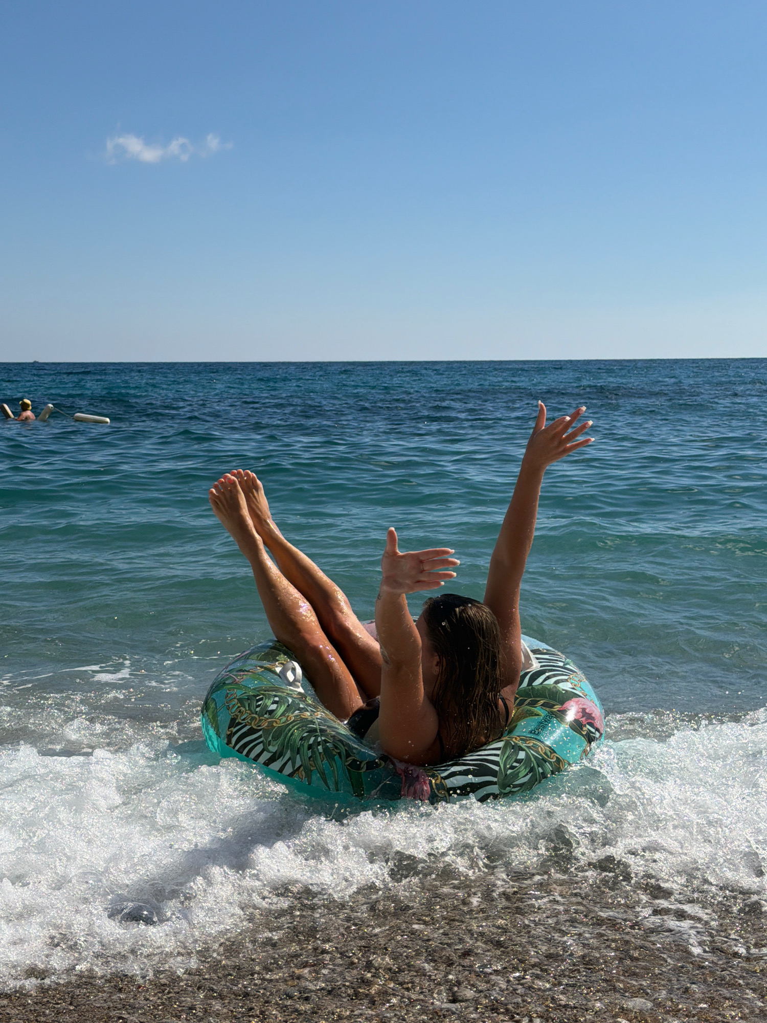 In Prijevor, Montenegro, on a clear day with a few wispy clouds, a person is seen from behind sitting in a patterned inflatable ring in the turquoise sea. The person's legs are extended upwards, out of the water, and their arms are raised in the air. White foam from a small wave surrounds the inflatable ring, which is decorated with tropical leaves and animal prints. The beach in the foreground is made of dark pebbles. In the background, the calm sea stretches to the horizon, and two other figures are visible in the water further offshore, appearing to be floating.
