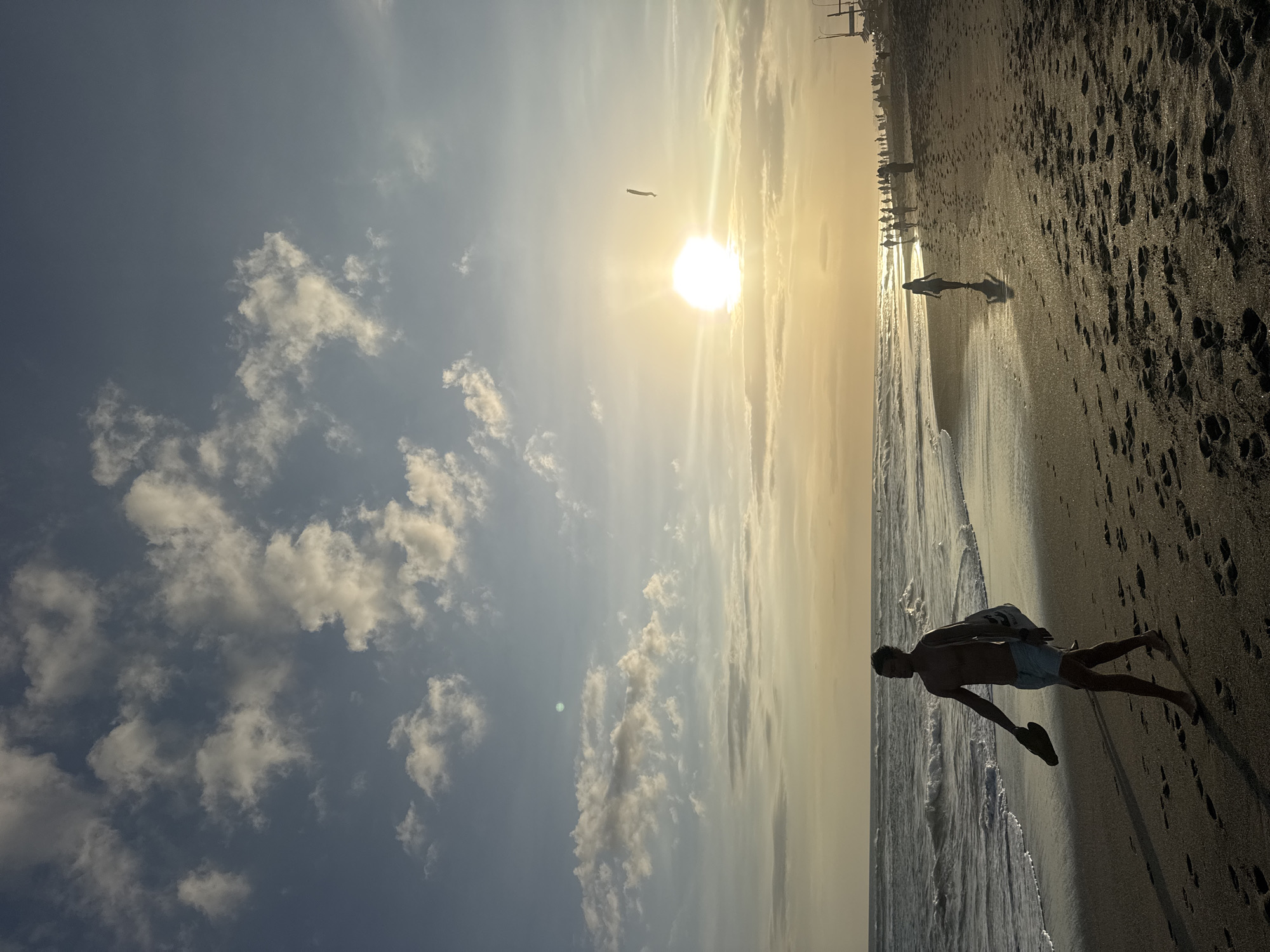 A man is walking on a beach in North Kuta, Indonesia. The sun is shining brightly, casting long shadows on the sand. The ocean waves are gently rolling onto the shore. Several other people can be seen in the distance, walking along the beach or sitting by the water. The sky is a mix of blue and white clouds, with the sun creating a hazy glow. The overall mood of the image is peaceful and relaxed, suggesting a beautiful day at the beach. The man in the foreground is silhouetted against the bright sun, making it difficult to discern his features, but he appears to be carrying a bag over his shoulder. The scene depicts a typical beach day in a tropical location.