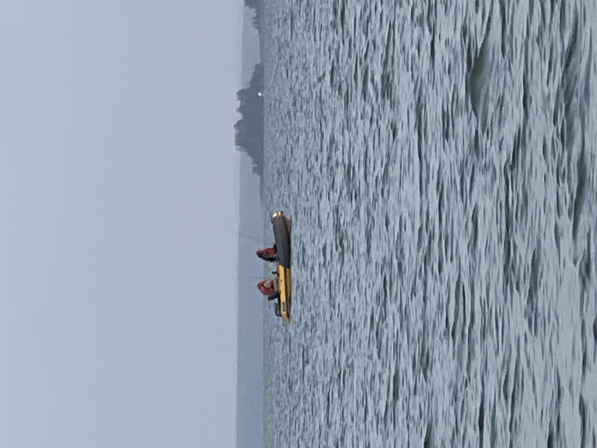 Two individuals are in a small yellow and gray boat on a large body of water. The boat is powered by an outboard motor. One person is wearing an orange life vest, and the other is wearing a red life vest. The water is choppy. In the background, there is a distant, tree-covered island and a hazy shoreline. The sky is overcast. The scene appears to be in Novopichugovo, Russia, which is known for its bodies of water and islands.