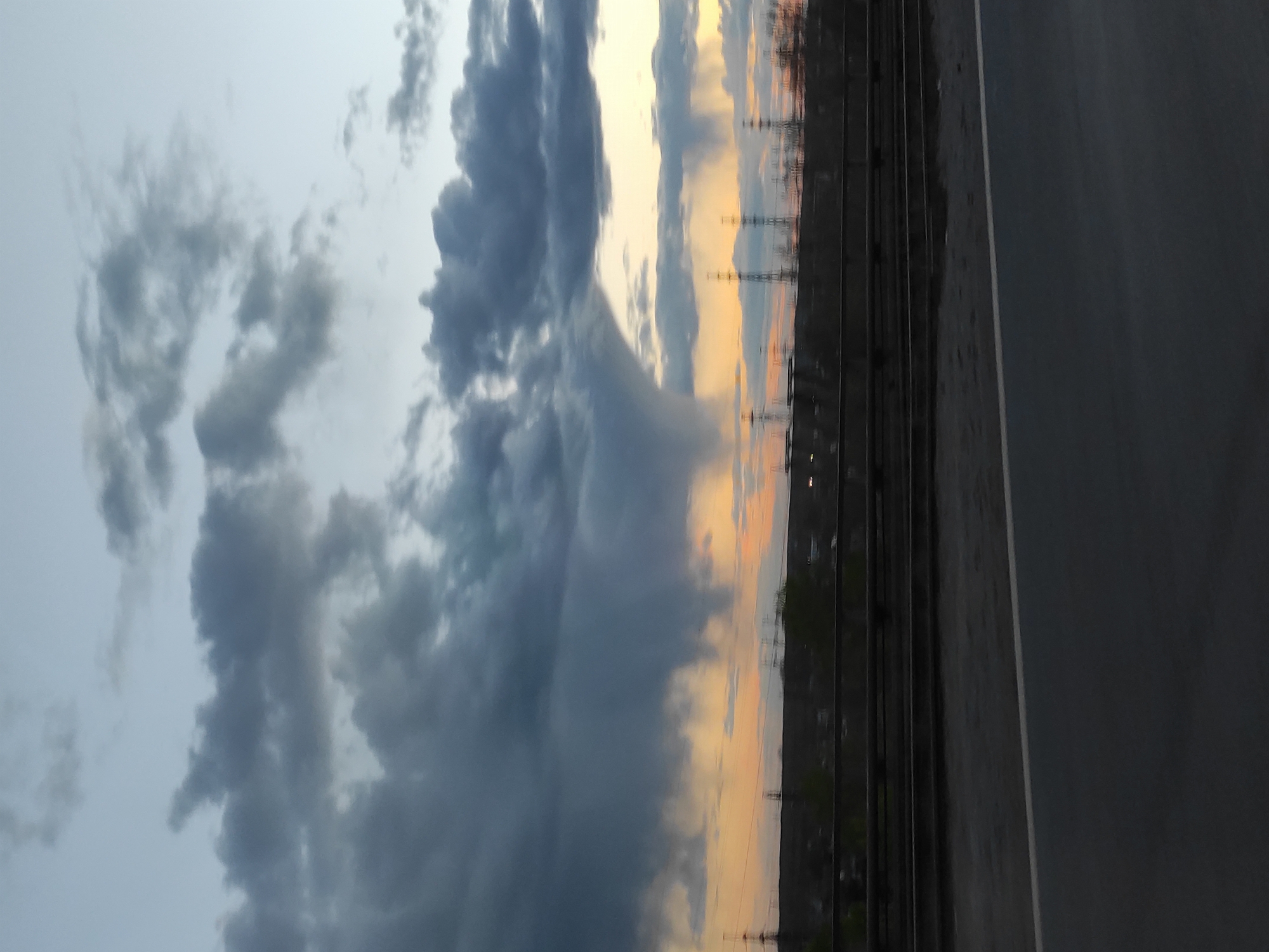 The image shows a view of the sky with dramatic, dark grey clouds dominating the left side. These clouds have a turbulent, almost tornado-like appearance in the center. To the right of the clouds, the sky transitions to a lighter, warmer color palette suggestive of either sunrise or sunset.
Below the sky, a landscape is visible. Power lines and towers are silhouetted against the colorful sky. In the foreground on the right side of the image, there is a grey asphalt surface, which appears to be a road or path. To the left of the road, a dark object is present.
Based on visual features such as the power lines and architecture, combined with the description, the location of this image is most likely Chaikovsky, Russia.
