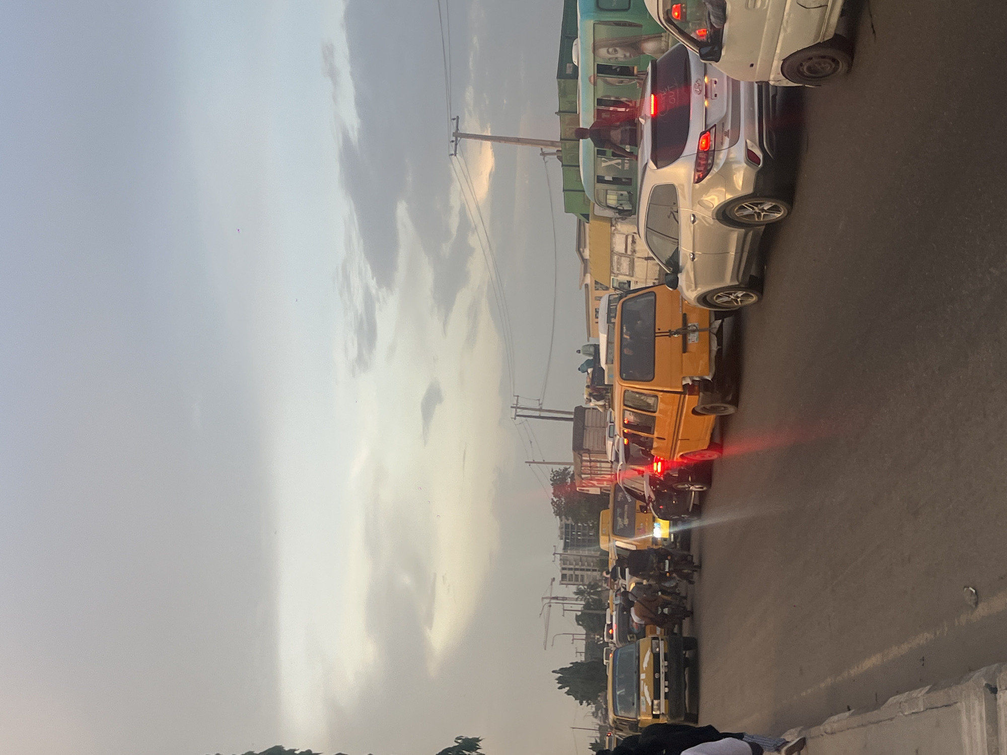 The image displays a congested street scene in Lagos, Nigeria, likely during late afternoon or early evening, given the diffused, warm light in the sky. The sky, occupying the left half of the frame, shows overcast clouds with lighter, golden-hued patches indicating the sun is low on the horizon.
Numerous vehicles are packed closely together on a multi-lane asphalt road, suggesting heavy traffic. The visible vehicles include several yellow public transport buses (commonly known as 'danfo'), smaller yellow tricycles ('keke napep'), and private cars, such as a silver Hyundai SUV. Many of these vehicles have their red brake lights illuminated. Motorcycles are also present, maneuvering between the larger vehicles.
Along the right side of the road, utility poles with overhead wires stand tall, and urban buildings are faintly visible in the background. One person is prominently seen standing on the rear of a green bus or truck, facing away from the viewer. The immediate foreground on the right features a low concrete barrier or wall. No discernible text is clearly visible on any of the vehicles or structures. The scene conveys a typical busy urban environment.