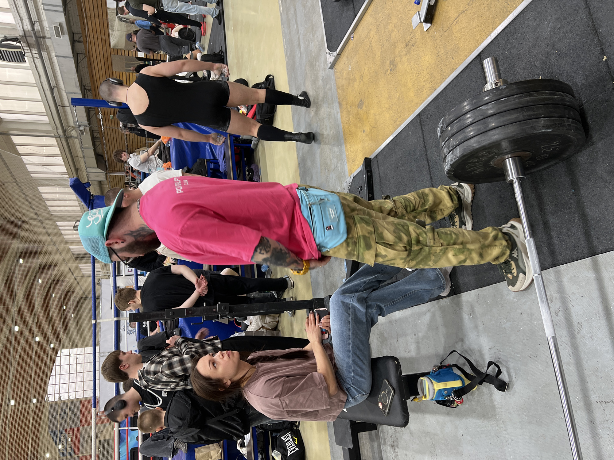 The image shows an indoor powerlifting competition in what appears to be Saint Petersburg, Russia. There are multiple individuals, mostly young adults, engaged in various activities. A woman wearing a brown shirt and jeans sits on a black bench, looking towards the scene. A man with a light blue baseball cap, a pink shirt, and camouflage pants stands near a barbell loaded with multiple weight plates. Other people are standing and sitting around the ring, some wearing athletic clothing. The overall atmosphere suggests a fitness event or competition. The venue appears to be an indoor gymnasium or sports facility.
