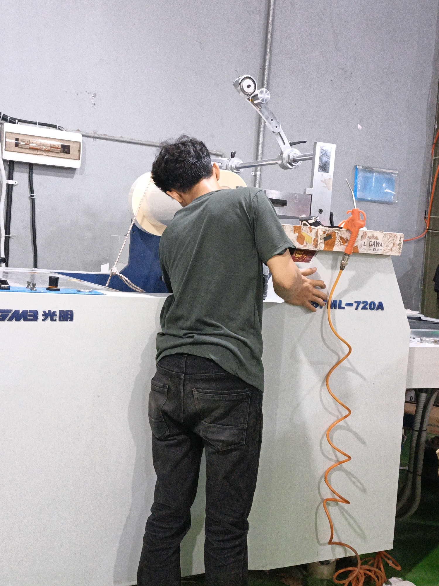 A male, appearing to be of Indonesian ethnicity, is working on a piece of industrial machinery in a brightly lit workshop. He is wearing a dark green t-shirt and dark wash denim jeans. The man is facing away from the camera, with his head and torso bent over the machinery, suggesting he is performing maintenance or operation.
The machinery is large and appears to be a cutting or grinding machine, with various metal components, including an articulated arm with a rotating head, and a large circular disc covered by a translucent shroud. A braided rope is attached to the shroud and secured to a part of the machine.
In the foreground, a white panel on the machine displays blue lettering that reads "GMB 光明" and "ML-720A". An orange air hose with a coiled shape is attached to the machine, suggesting a pneumatic system is in use.
To the left of the machinery, a white electrical distribution box with multiple switches is mounted on a grey wall. The environment appears to be a manufacturing or repair facility, with concrete walls and industrial equipment. The floor is a dark color, possibly concrete or tile.
The overall scene depicts a worker engaged in a task within an industrial setting. The location is likely North Jakarta, Indonesia, given the presence of the brand "GMB Guang Ming", which is a known manufacturer of industrial equipment in Indonesia.