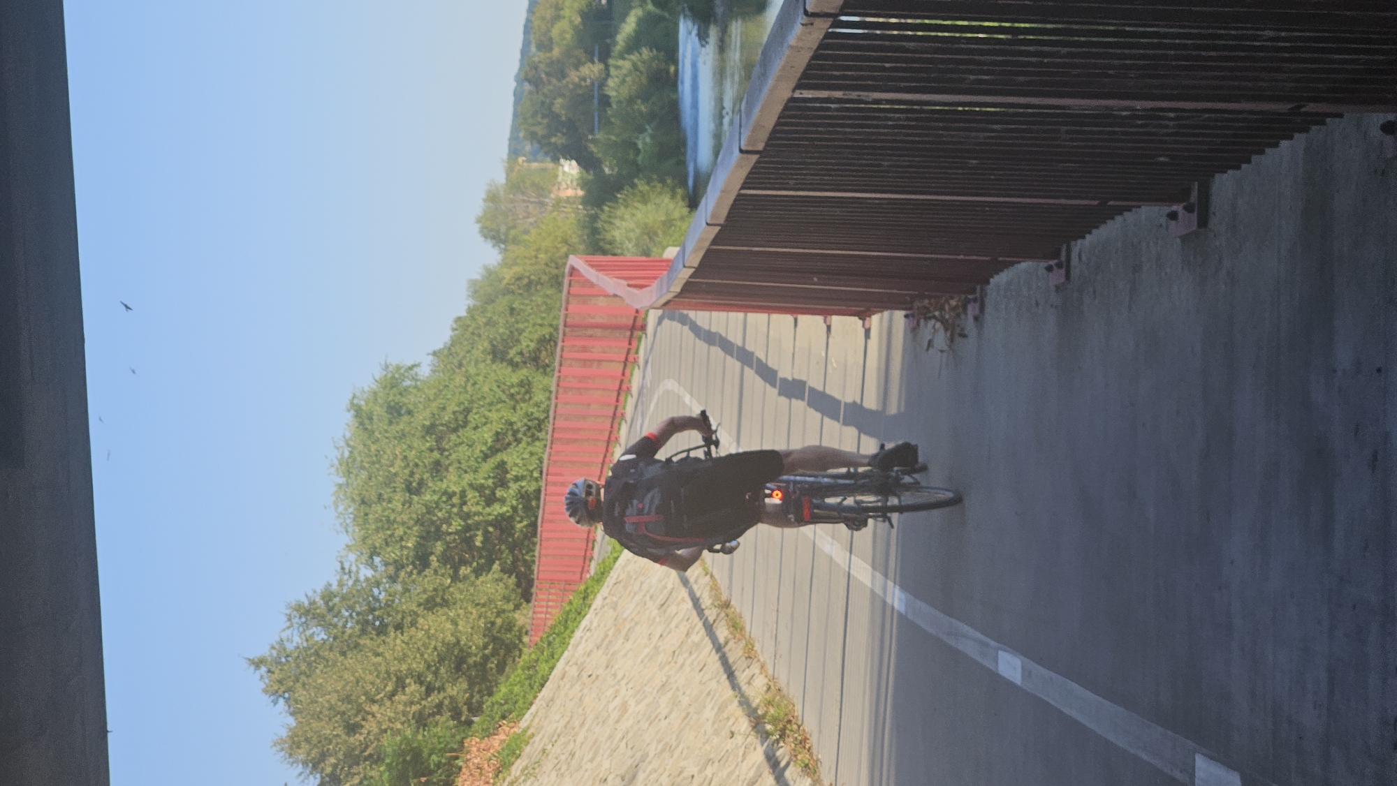 A lone cyclist is riding a bicycle on a paved path next to a red metal fence. The cyclist is wearing a helmet and a backpack. The path appears to be a dedicated bike lane, separated from traffic. In the background, there is a body of water, possibly a river, with trees and buildings along its banks. The sky is clear and blue, and the sun is shining. The image captures a peaceful scene of outdoor recreation in a city park or along a waterfront. This location is the cycling path along the Torysa river in Prešov, Slovakia.