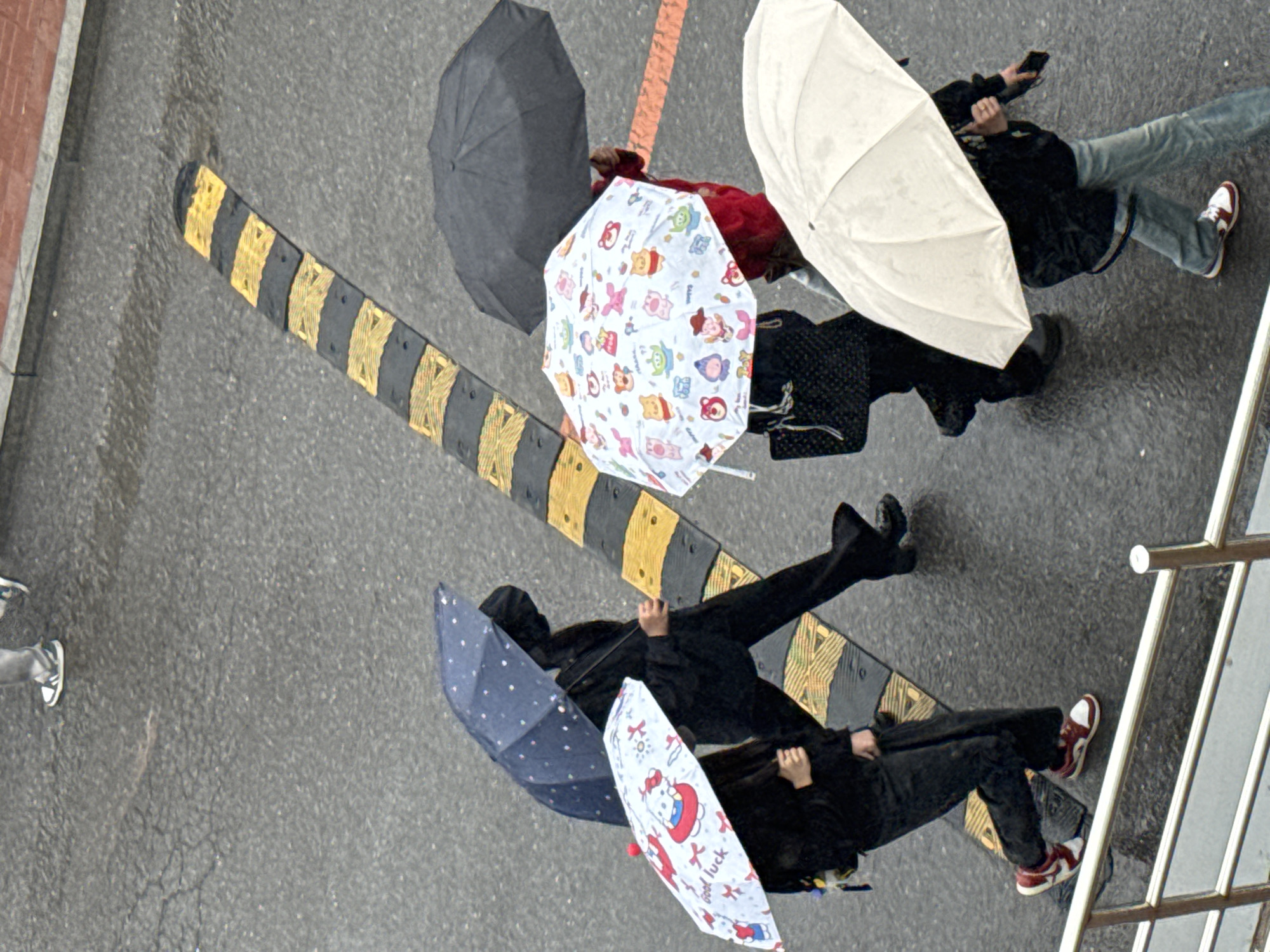 The image, taken from an elevated perspective, depicts a rain-soaked street scene, possibly in Zhongtun, China. Six individuals are visible, each holding an umbrella. There is a dark grey umbrella, a white umbrella, an umbrella with cartoon figures, a blue polka-dotted umbrella, and a white umbrella with a red cartoon. The people are walking in the rain. A yellow and black striped traffic calming device is present on the road surface. The road is asphalt. A small section of a metal railing is visible at the bottom right corner. The overall atmosphere is wet and overcast.