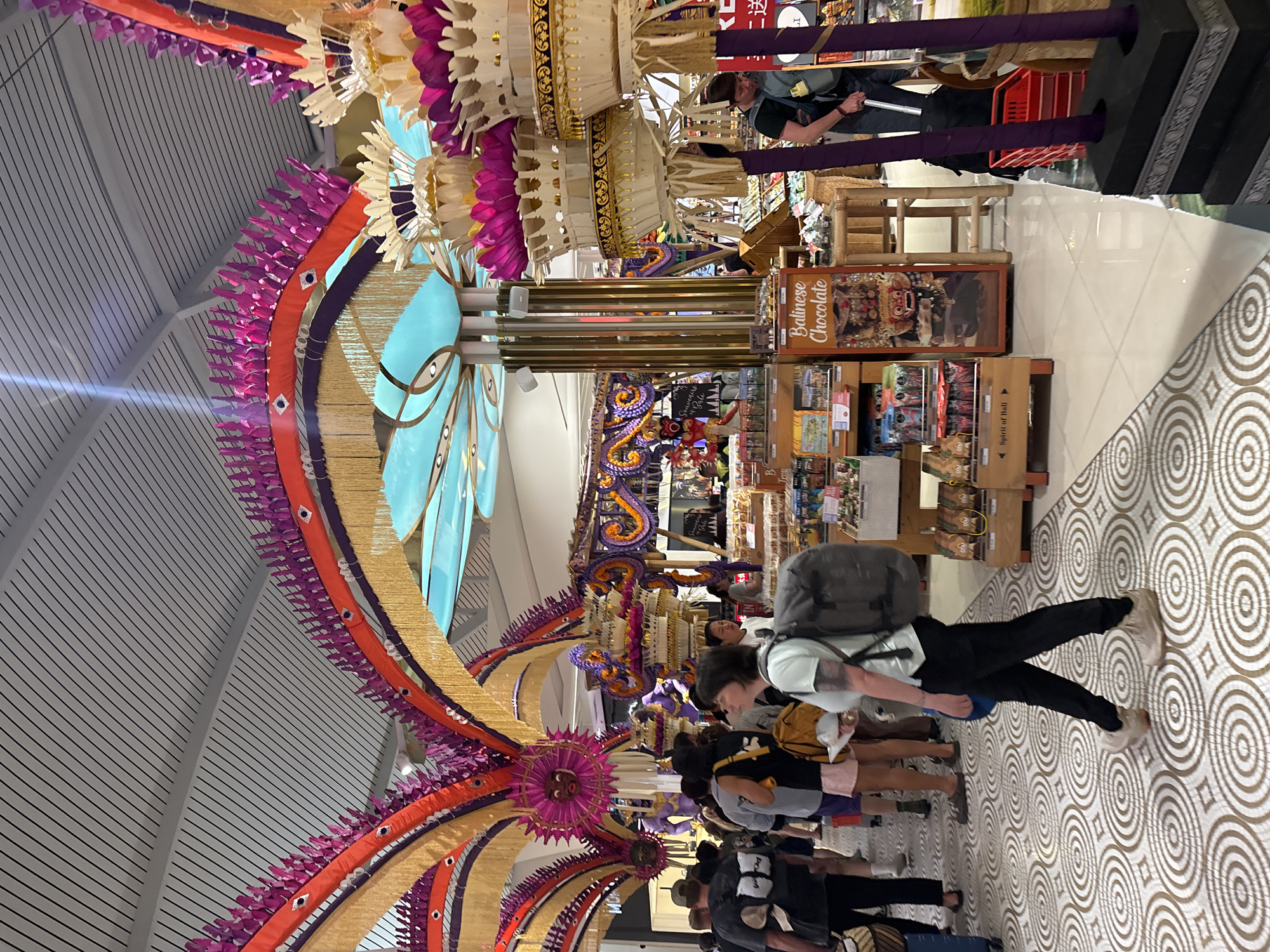 The image shows the interior of an airport, likely the Ngurah Rai International Airport, in Kuta, Bali, Indonesia. The ceiling is decorated with ornate, colorful, symmetrical floral and other traditional Balinese decorations that hang down. There are people walking through the airport; some are walking toward the camera, and others are walking away. A person with a backpack and wearing a light-colored shirt and black pants is walking toward the camera. There is a shop on the right side of the image with various items for sale, including "Balinese Chocolate." The floor has a patterned tile design. A portion of the ceiling is visible, featuring a striped pattern.