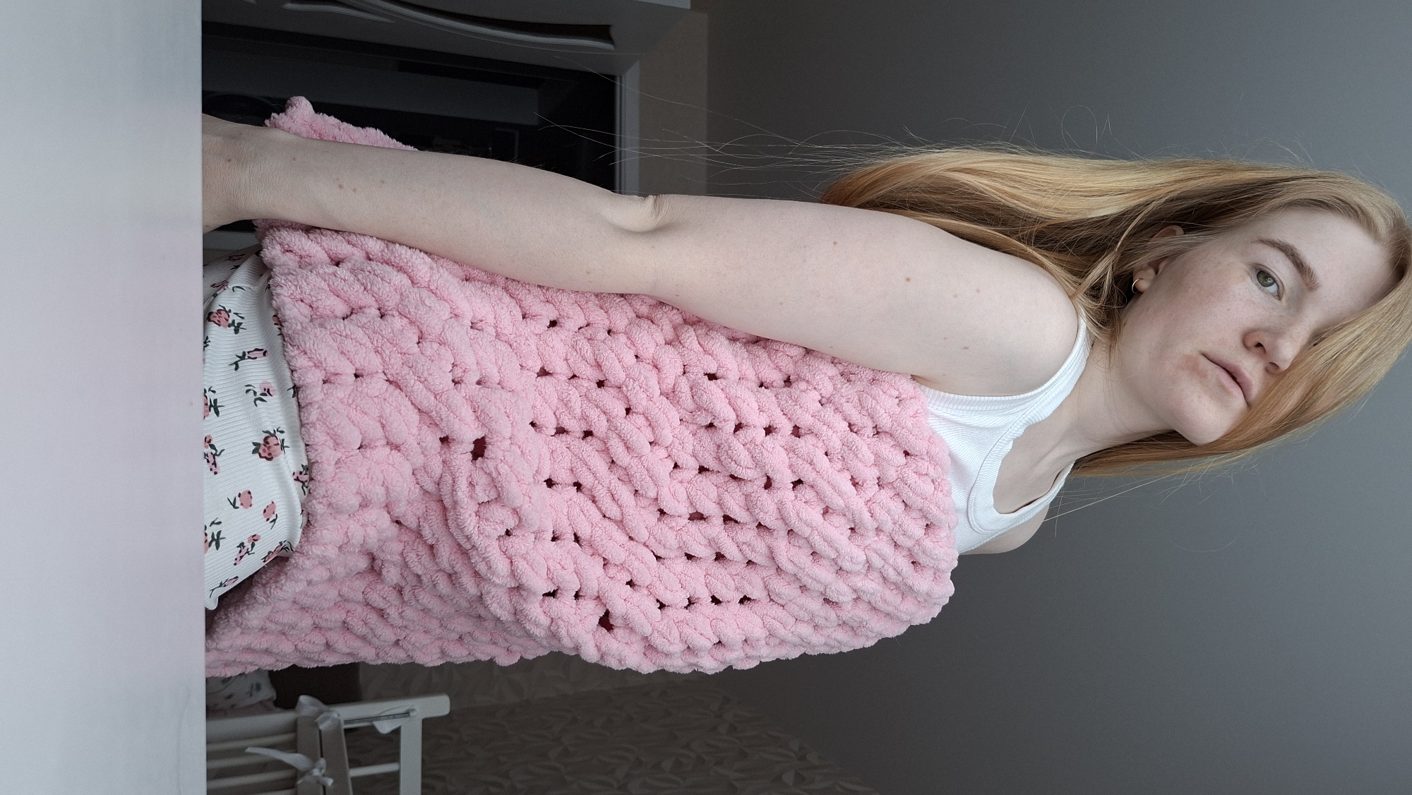 The image depicts a fair-skinned woman with freckles and strawberry-blonde hair, posing indoors with a light pink, chunky knit blanket. She wears a white tank top and floral-patterned pajama bottoms. The environment suggests a domestic setting, with a white wall and a gray wall visible in the background. A portion of a cabinet or appliance is visible behind her left shoulder. A crib is partially seen at the lower left of the frame. There is no verifiable information to suggest the location is Chaikovsky, Russia.