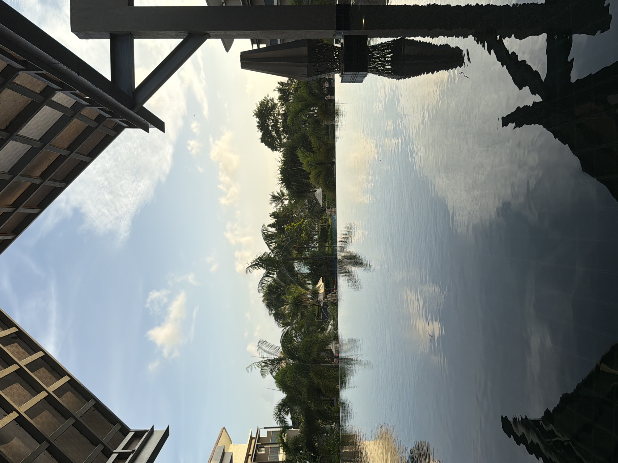 This image depicts an infinity pool at what appears to be a resort or hotel in the city of Bang Muang, Thailand. The pool's surface is calm, reflecting the sky and surrounding greenery. In the background, lush tropical trees and palms line the horizon, with a glimpse of a modern building on the left. A distinctive, angular dark sculpture or lamp stands on the right edge of the pool, casting a reflection on the water. The architectural elements framing the top of the image suggest a covered walkway or pavilion, with exposed wooden planks and metal beams against a pale blue sky dotted with wispy clouds. The lighting indicates it is likely late afternoon or early evening. No people are clearly identifiable or engaged in any specific actions within the frame.