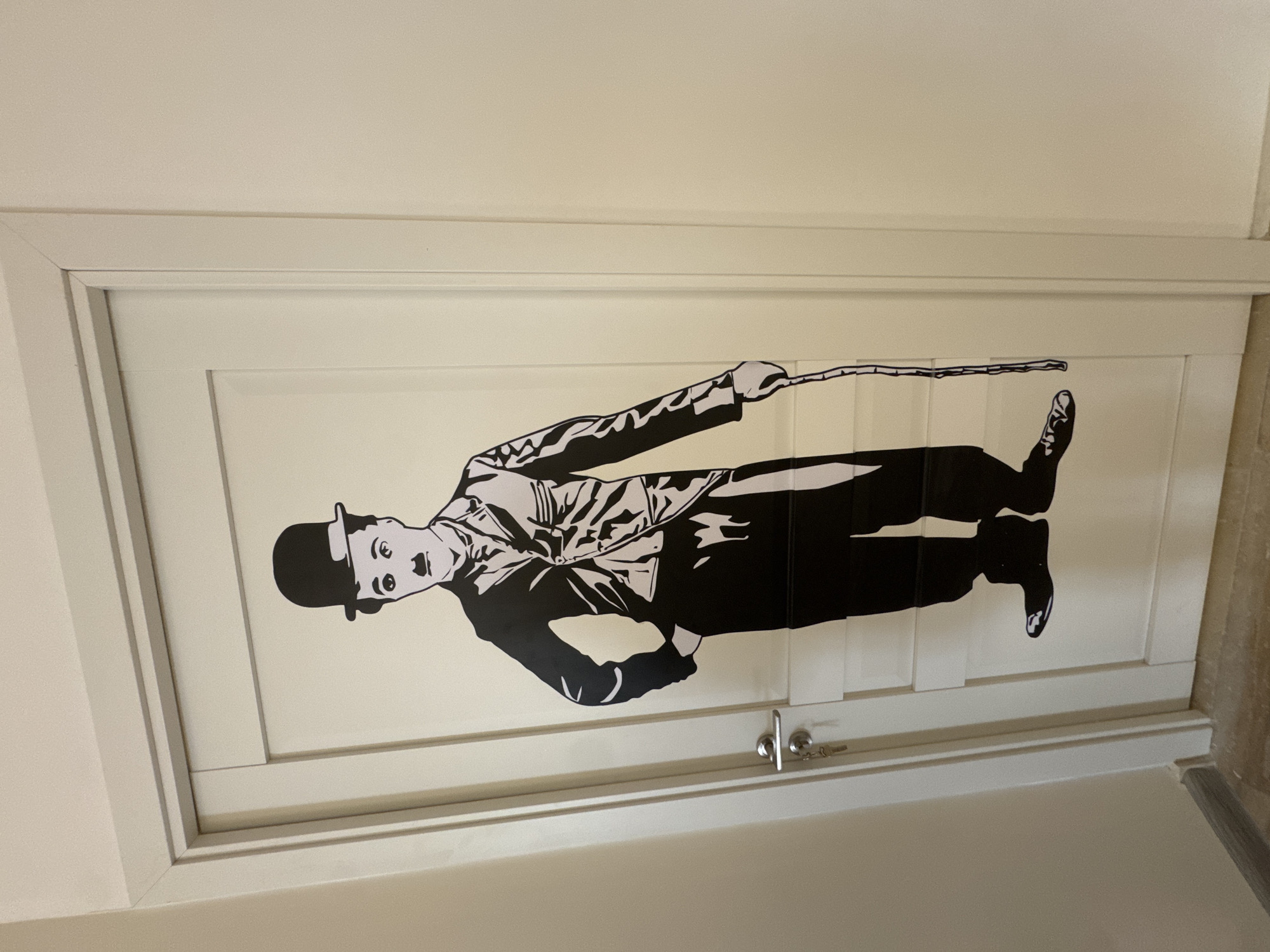 The image presents a vertical view of a white interior door featuring a large, black and white graphic of Charlie Chaplin in his iconic "The Tramp" persona. The high-contrast graphic dominates the door, showing Chaplin standing, holding a cane in his right hand and with his left hand on his hip. He is depicted with his characteristic bowler hat, small mustache, and expressive face, looking towards the viewer.
The door is painted white and appears to have recessed paneling. A simple, chrome-colored door handle and a keyhole with a key inserted are visible at the bottom right portion of the door. The door is set within a white frame, surrounded by light-colored walls, indicating an indoor setting. No specific activity is occurring, as the image focuses on the static decorative element. No text is visible in the photograph. The context provided indicates this location is in Yerevan, Armenia.