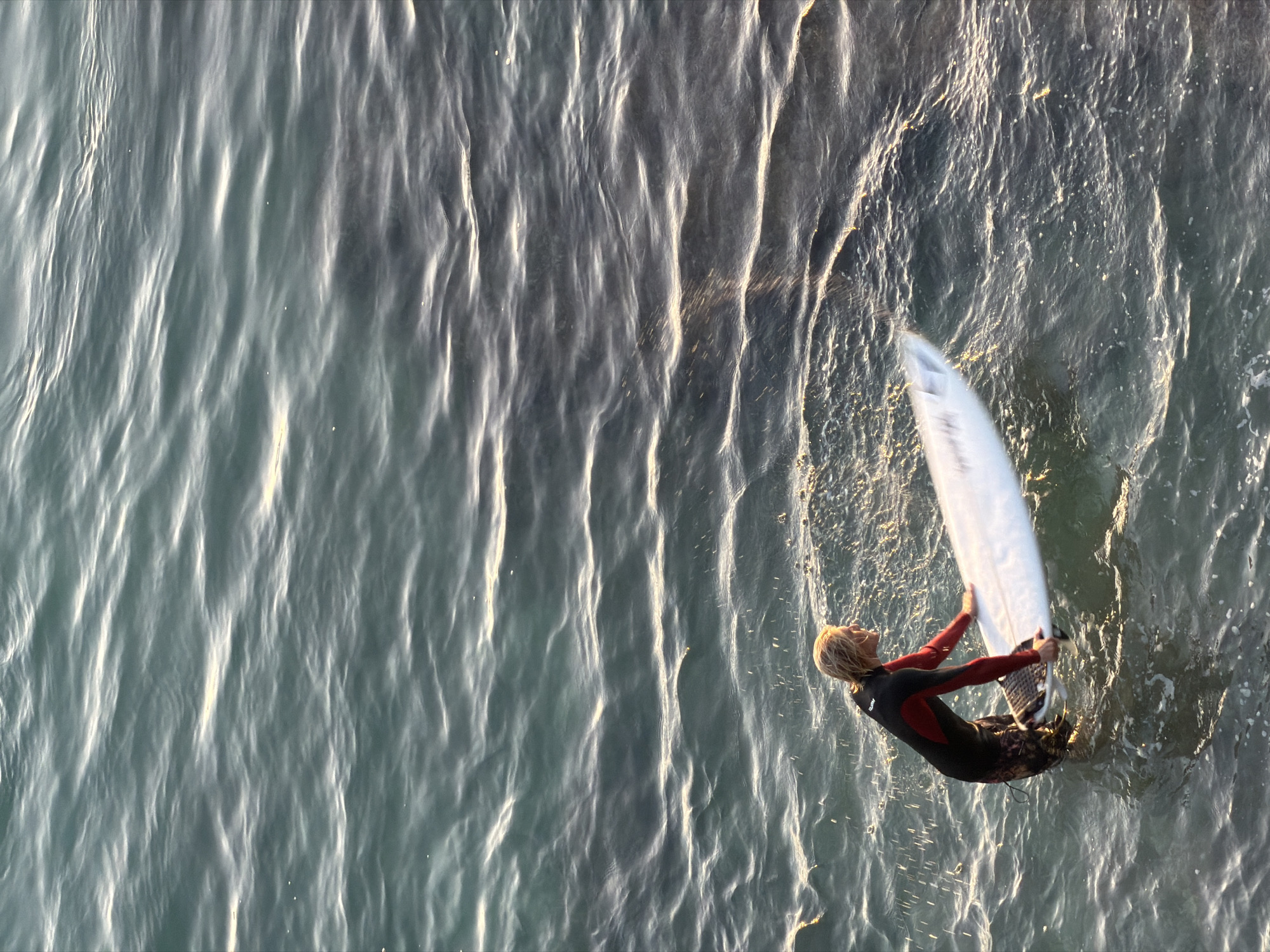 A surfer in a black and red wetsuit stands in choppy, sunlit water with a white surfboard. The surfer's blond hair is visible as they look to the right. The water is a dark teal with whitecaps and reflections of the sun. The image appears to be taken from an elevated viewpoint looking down at the surfer. The location is the coast of Kuta Selatan, Indonesia, known for its surf breaks.