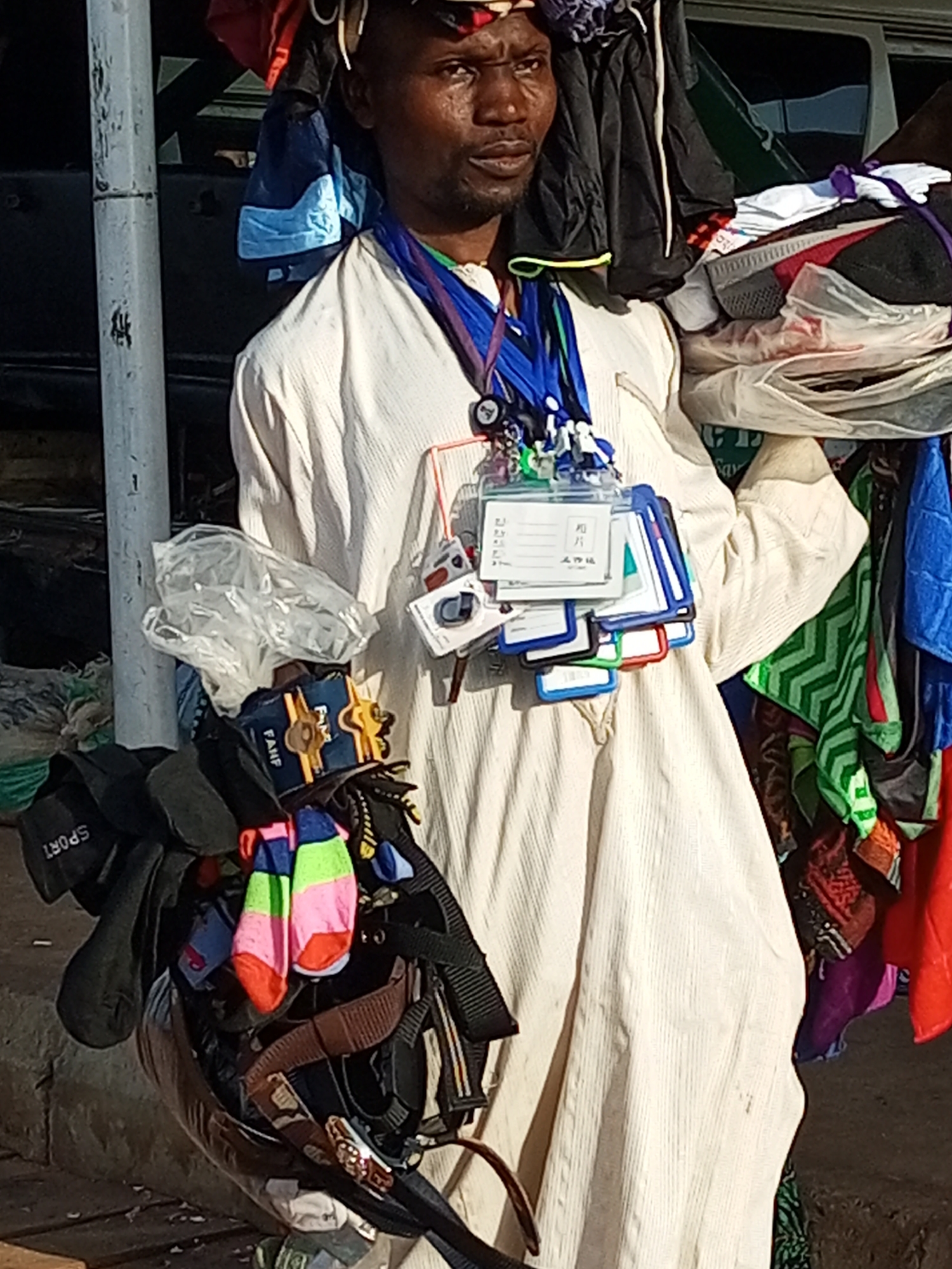 The image shows a man standing outdoors in what appears to be a busy marketplace in Gombe, Nigeria. He is wearing a light-colored, striped robe and has a collection of items hanging from lanyards around his neck. These items include several ID cards, some small figures, and what look like small electronic devices. He is also carrying a large bundle of folded clothing and textiles over his right shoulder. To his left, a collection of hats, socks, and what appear to be straps or belts are attached to a dark object, possibly a cart or display. Behind him, colorful fabrics, including a green chevron pattern, are visible. The environment suggests an informal street vending setting.