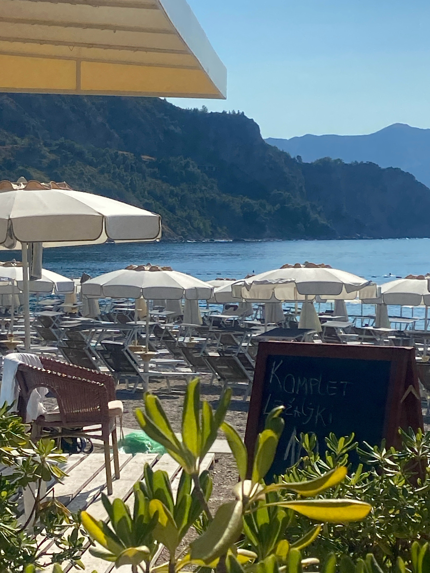 The image depicts a sunny day at a beach in Prijevor, Montenegro. In the foreground, lush green foliage partially obscures a wooden walkway. Beyond the walkway, numerous beach chairs and umbrellas are arranged on the sand, indicating a well-maintained resort or public beach. The umbrellas are mostly white, providing shade over the empty chairs. In the middle ground, the calm blue sea stretches out, with a few people visible swimming or floating in the water. The background is dominated by a range of majestic, verdant mountains that slope down to meet the sea, with the clear blue sky above. A dark chalkboard sign with white lettering is visible in the lower right portion of the image, displaying the text "KOMPLET LEZALJKI". This suggests the beach offers services related to the rental of beach loungers. The scene evokes a sense of tranquility and relaxation, characteristic of a Mediterranean vacation spot.