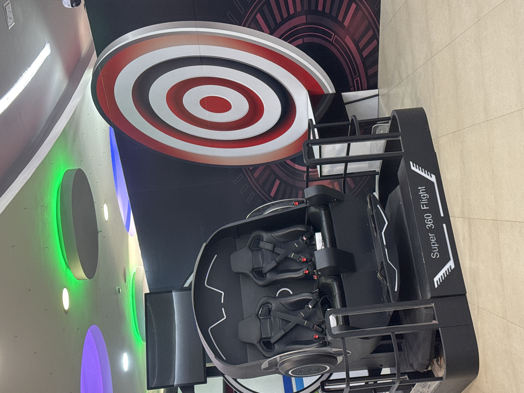 This image captures an indoor entertainment setting in Yerevan, Armenia, featuring a "Super 360 Flight" virtual reality or motion simulator ride. The main subject is a black, futuristic-looking apparatus designed for two passengers, each seat equipped with substantial safety harnesses. The name "Super 360 Flight" is prominently displayed in white lettering on the side of the ride, suggesting its interactive and rotational nature.
The background elements contribute to a dynamic and modern atmosphere. Behind the ride, a large, multi-layered circular target design, featuring concentric rings of red, white, and grey, is affixed to a dark wall that also displays subtle, dark red geometric patterns. To the left, another wall is adorned with circular cutouts, illuminated by vibrant green and purple LED lights, enhancing the entertainment venue's ambiance. The floor is covered with light-colored, possibly beige, tiles. A small security camera is visible in the upper-left corner of the frame. No individuals are present, indicating the ride is currently unoccupied.