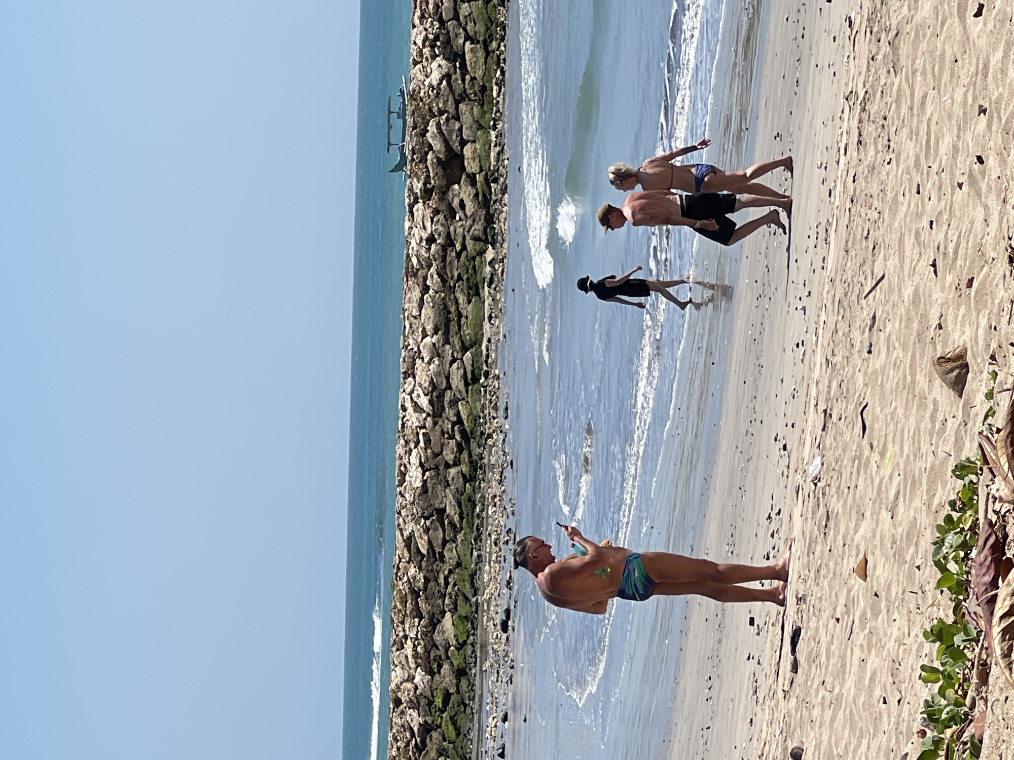 A group of people are walking on a beach in Kuta, Indonesia. In the foreground, a man in swimming trunks looks at his phone. Behind him, a couple walks along the water's edge. Further back, a person in a black dress walks towards the water. In the background, waves crash against a rocky barrier. The sky is clear and blue.