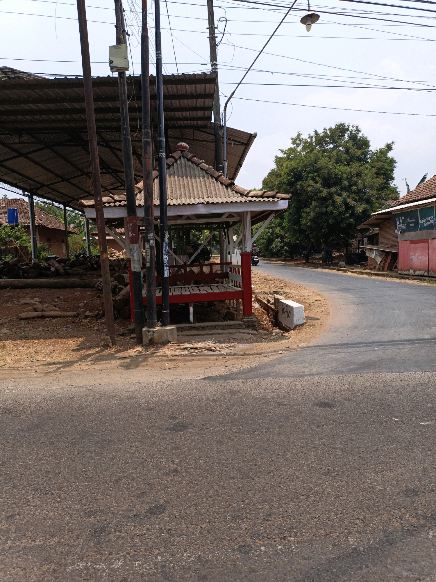 The image shows a street scene in Cikal, Indonesia, featuring a small, open-air pavilion with a red base and white roof. The pavilion is situated on the side of a paved road, which curves to the right. Scattered around the pavilion are several logs of wood. Behind the pavilion, a larger, covered structure with a corrugated roof is visible, supported by wooden poles. To the right of the road, a building with a red facade and signs advertising "Beda Masa Baru" is present. A single motorcyclist is seen riding on the road. Overhead, numerous power lines crisscross the sky. The environment appears rural, with trees and vegetation surrounding the area. The sky is overcast with a few clouds.