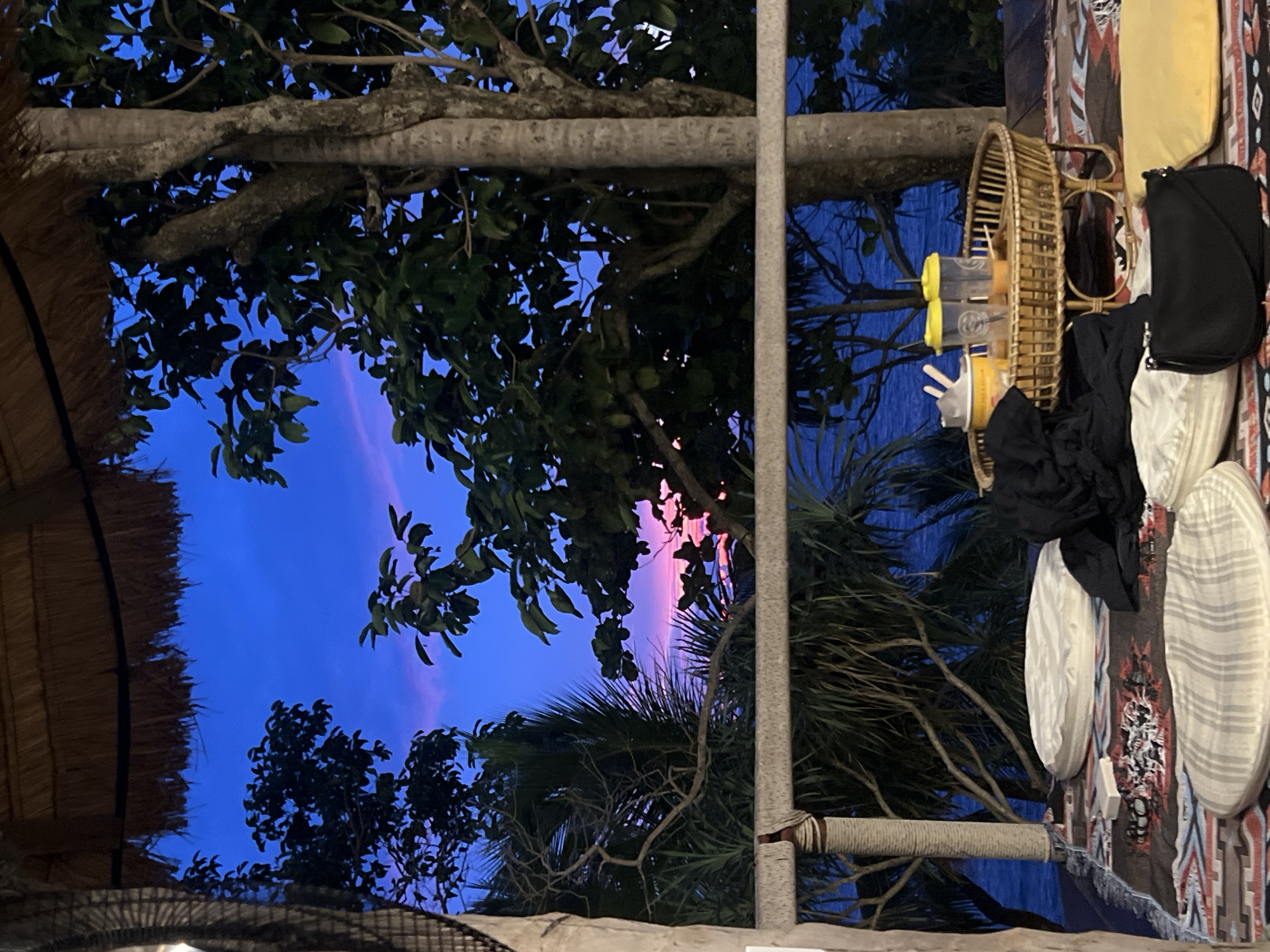 The image depicts an outdoor setting at dusk, likely in Karon, Thailand, given the location context. The sky is a deep blue, transitioning to a soft pink and purple hue near the horizon, suggesting a sunset or twilight. Silhouetted against this vibrant sky are the dark green leaves and branches of tropical trees, including what appears to be a palm tree. A thatched roof structure is visible on the left, hinting at a beachside cafe or resort.
In the foreground, several objects are arranged on what looks like a patterned rug or mat. These include a woven basket filled with several cups containing yellow liquid and straws, possibly drinks. Adjacent to the basket are sunglasses and a black bag. Other items of clothing, including black and white fabric, are also present. A sturdy wooden pole wrapped with rope stands vertically in the middle of the frame, dividing the scene. The overall mood is relaxed and serene, characteristic of a tropical evening. There is no visible text in the image.