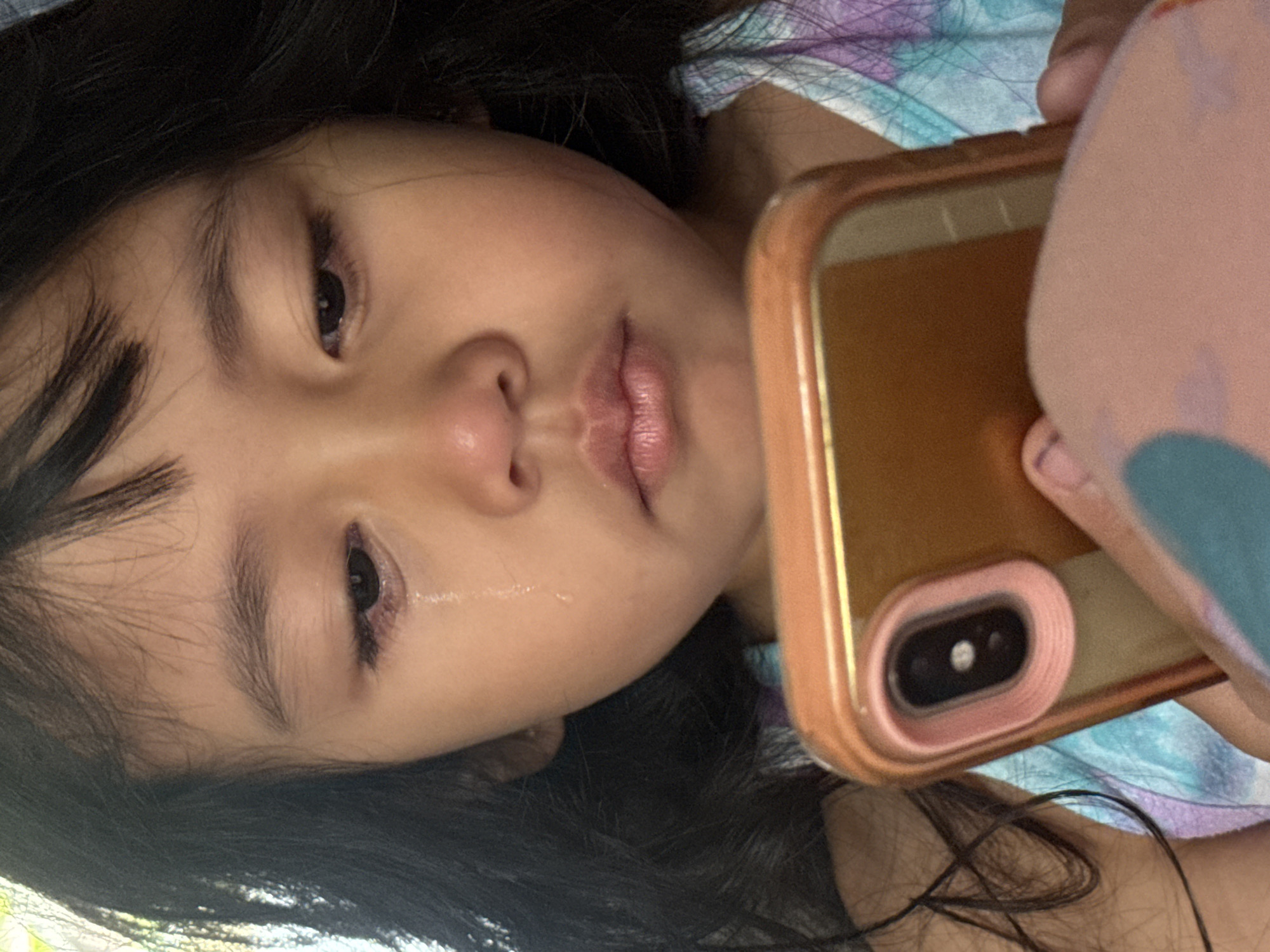 A young girl, appearing to be of Southeast Asian descent, is crying and looking at a smartphone. She has dark hair and is wearing a tie-dye shirt. The phone is gold with a pink case and is held in her hand. The background is blurred but suggests an indoor setting. There are no identifiable landmarks or features that can place this image specifically within Davao City, Philippines.