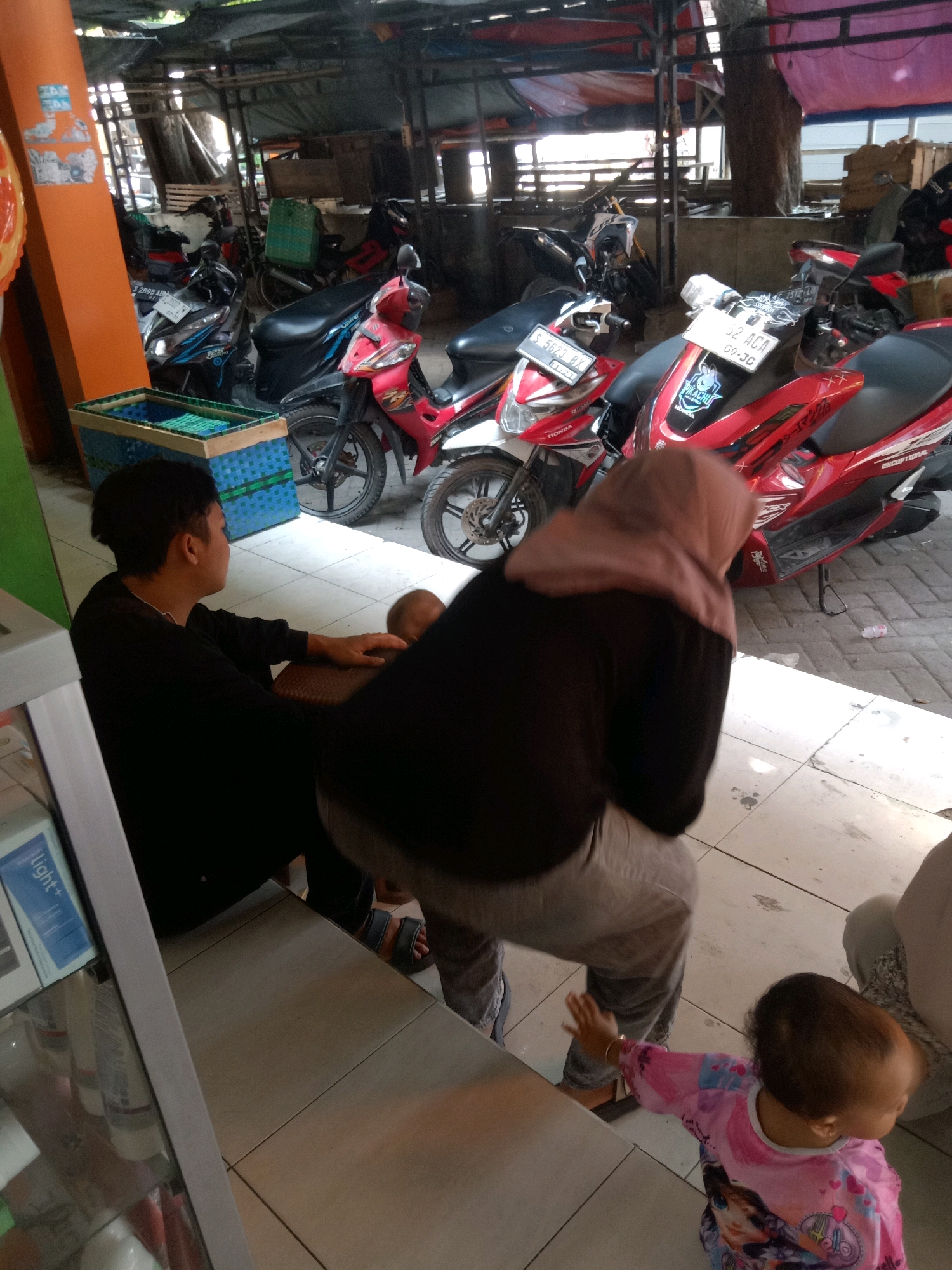 The image depicts a scene in Sugihwaras, Indonesia, likely at a market or roadside stall. The foreground features a young child in a pink shirt, reaching out their hand. To the left, a young man in a black shirt is seated, looking away from the camera. Behind them, a person wearing a hijab is seated, facing away and appearing to be tending to something. In the background, several motorcycles are parked, some with red and white detailing, and one appears to be a scooter. A blue and green woven basket is visible on the left side of the image. The environment suggests an open-air setting, with a corrugated roof overhead and tiled flooring.