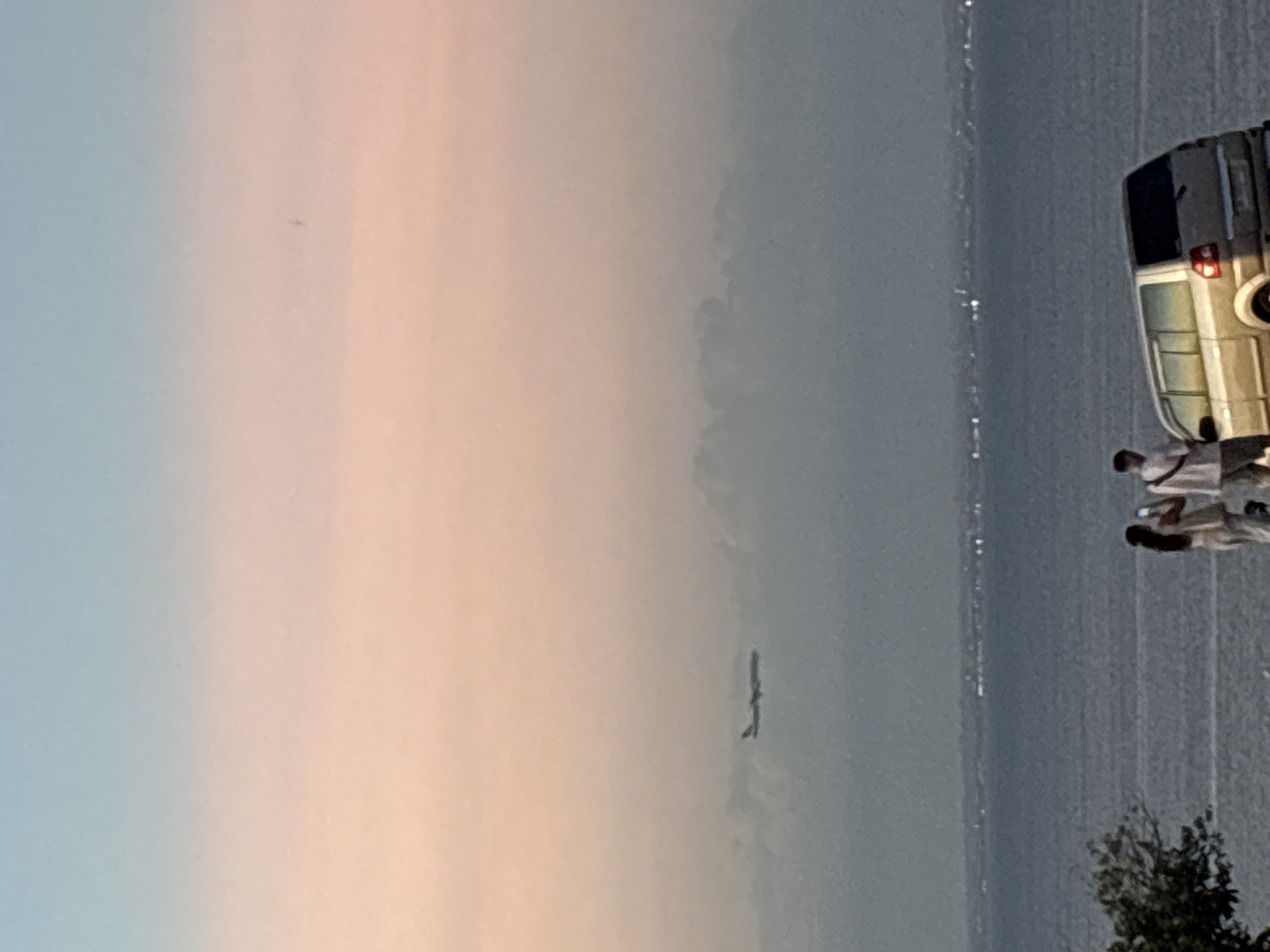 The image shows a horizontal view of the coastline in Kuta Selatan, Indonesia. In the foreground, a vehicle and two individuals stand on the right side of the frame. The background reveals a hazy horizon line where the sky meets the sea. The sky exhibits a gradient from light blue to peach. The coastline is slightly illuminated with faint city lights.