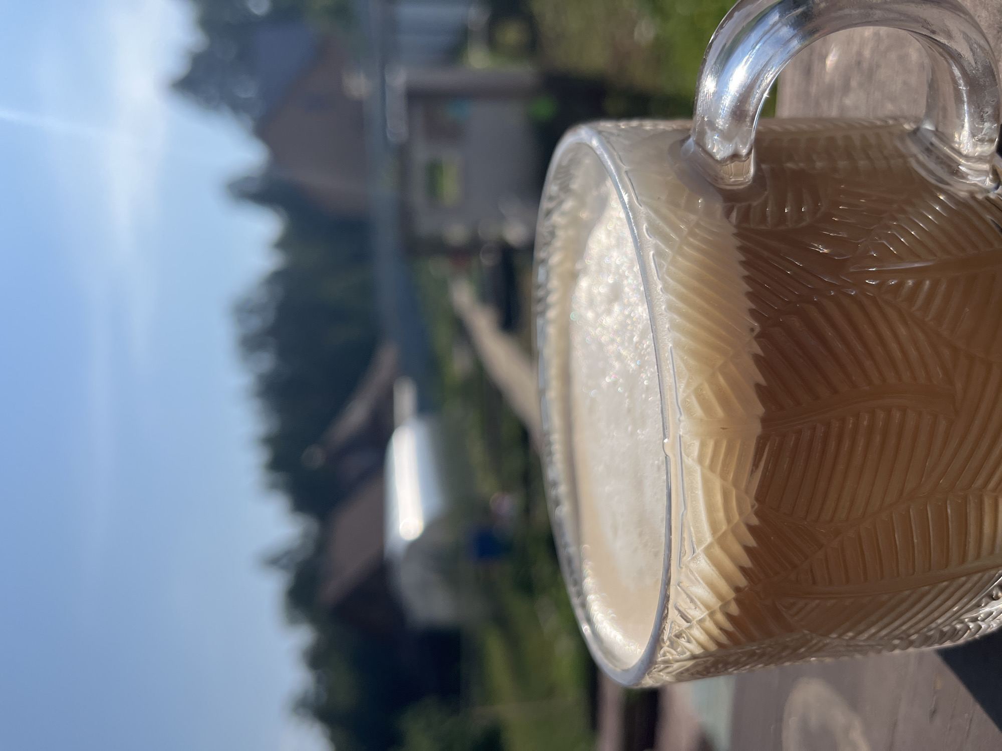 An outdoor shot shows a glass mug filled with a frothy, layered beverage, likely coffee, placed on a wooden surface. The mug has a textured pattern of diagonal lines. In the blurred background, several wooden houses with red roofs are visible, along with a greenhouse and trees under a clear blue sky. The setting suggests a dacha or country house environment, potentially in Chaikovsky, Russia, given the commonality of such residences in the region. The sun is shining, casting shadows and highlighting the sparkle on the drink's surface. There are no people or animals visible in the frame.