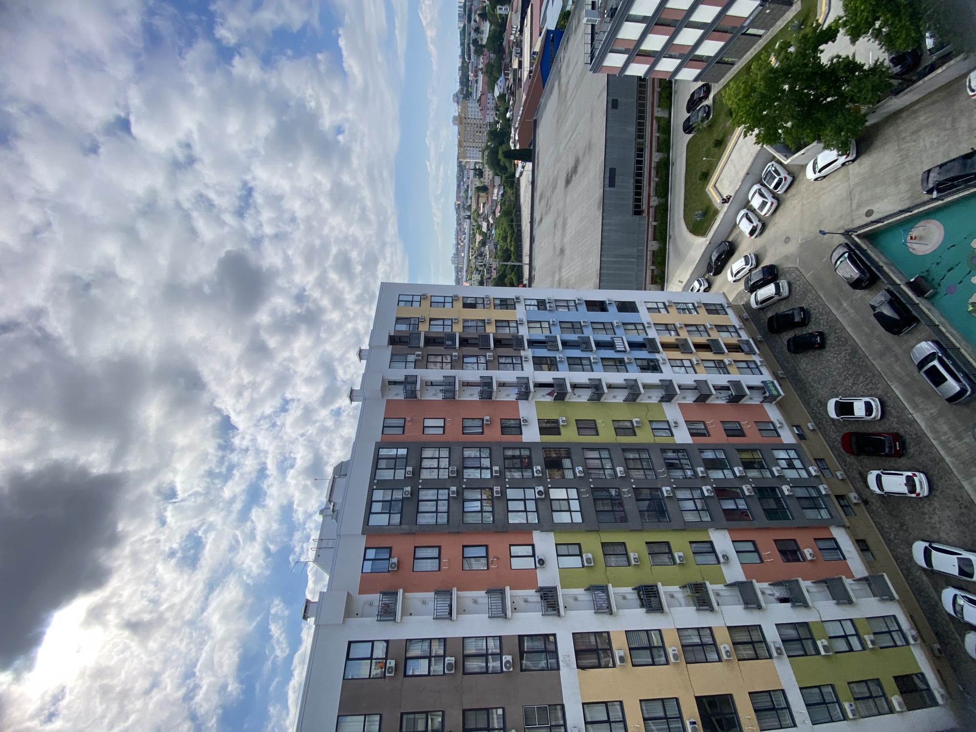 In Sochi, Russia, a modern, multi-colored apartment building with balconies and air conditioning units dominates the foreground under a partly cloudy sky. Below the building, a parking lot contains several parked cars, including white sedans and SUVs, black cars, and a dark red car. Adjacent to the parking lot is a playground area with blue and green surfaces. Further in the background, a large, flat-roofed industrial building is visible, along with other urban structures and distant green hills. The scene captures a residential area on a day with a mix of sun and clouds.