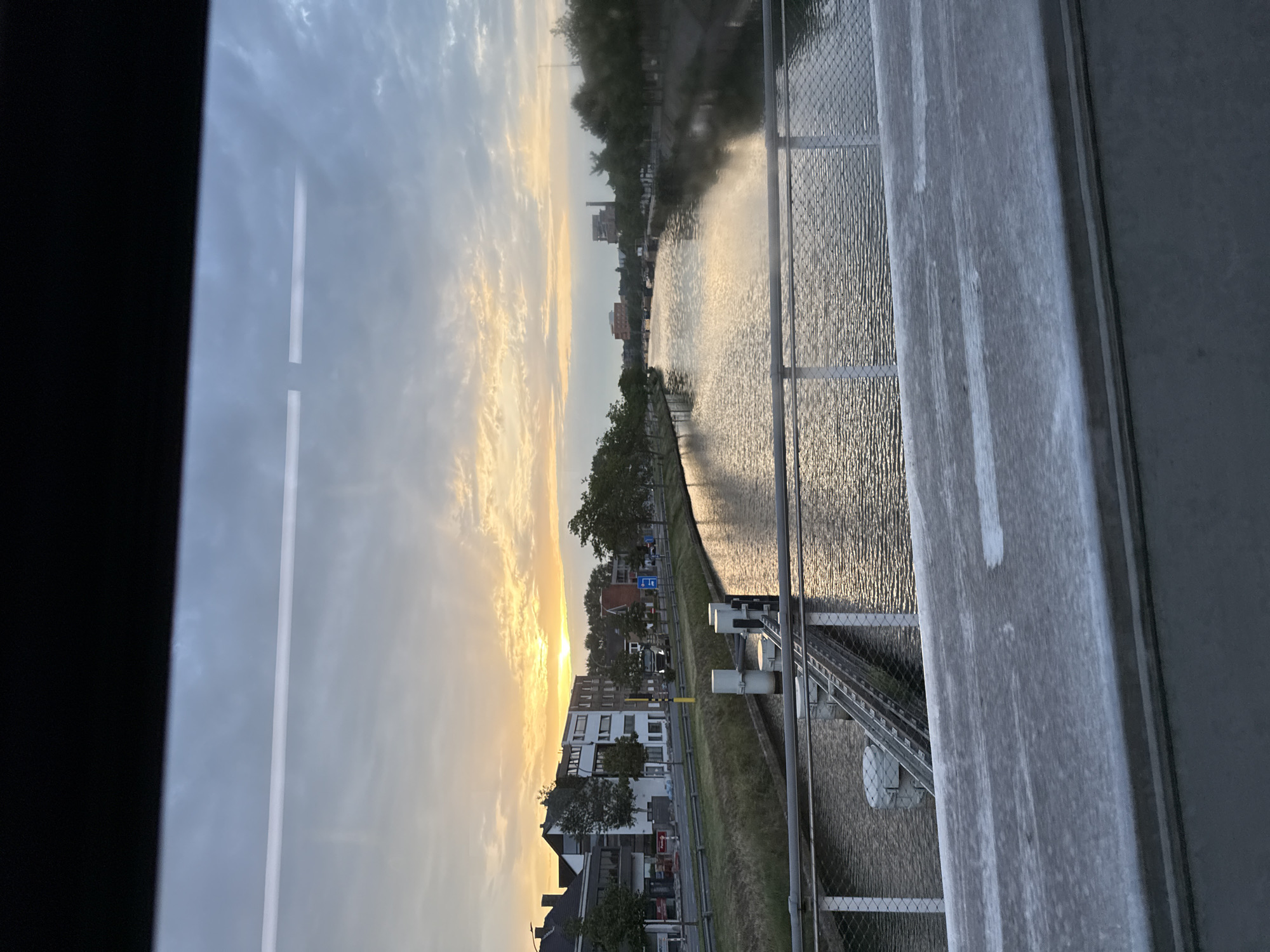 This is a full shot, eye-level, outdoor, medium distance photograph taken on a cloudy day. The sun is setting, casting a warm golden glow on the sky and reflecting on the water. In the foreground, a concrete barrier with some white markings is visible. Beyond the barrier, a metal railing with a mesh infill separates the viewer from a body of water. On the left side of the water, a canal or river runs parallel to a road lined with buildings and trees. The buildings appear to be residential or commercial, with some signs of construction. On the right side of the water, more trees line the bank, with some industrial buildings or structures visible in the distance. The water itself is calm, with ripples reflecting the sky and the surrounding environment. The scene depicts an urban waterway with a bridge in the foreground, overlooking the sunset. The location is identified as Bruges, Belgium.
