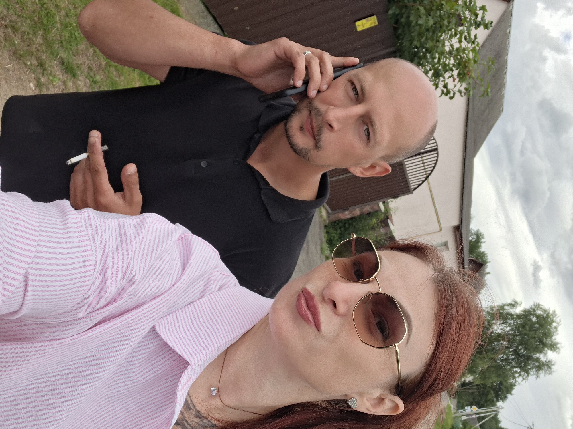 This is a selfie taken outdoors with a man and a woman. The man, on the left, is bald with a goatee and is wearing a black polo shirt. He is holding a mobile phone to his ear and has a cigarette in his hand. The woman, on the right, has red hair and is wearing a pink and white striped shirt and sunglasses. She is puckering her lips for the selfie. In the background, there is a fence, a gate, a building, and trees, suggesting a residential area. The location is identified as the city of Myadzyel, Belarus.