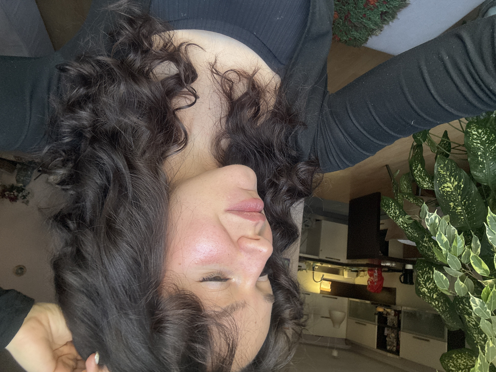 A portrait of a woman with dark curly hair wearing a black long-sleeved shirt is rotated 90 degrees counter-clockwise. Her eyes are mostly closed, and her face is slightly flushed. The background reveals a domestic setting: kitchen cabinets, countertops with objects on them, and houseplants (possibly a Dieffenbachia or a similar variegated plant). The scene is not identifiable with Burgas, Bulgaria, without additional evidence.