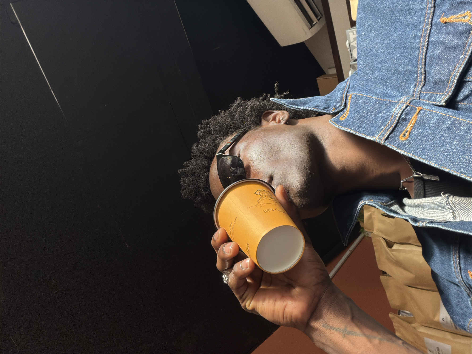 An image taken from a low-angle perspective, rotated 90 degrees clockwise, depicts an adult individual, likely male, with dark skin and short, curly dark hair. The person is wearing dark sunglasses pushed up on their nose, a blue denim jacket with prominent orange stitching, and a silver pendant necklace. A tattoo is visible on their forearm, and a ring is worn on one finger.
The individual is holding an orange paper coffee cup, which obscures their mouth, suggesting they are in the act of drinking. Bright sunlight illuminates the person's face and jacket, indicating daytime. In the background, a large dark wall or surface dominates the left side, featuring a thin, bright diagonal line. A white air conditioning unit or vent is visible in the upper right. A brown paper bag is partially visible tucked behind the person's jacket on the right.
The setting appears to be an indoor or sheltered outdoor space, potentially a cafe or shop in London, United Kingdom. Visible text on the coffee cup includes "100% C...", "coffee.com", and "www.green...", along with a graphic of a winged creature. The person's specific emotion is not discernible.