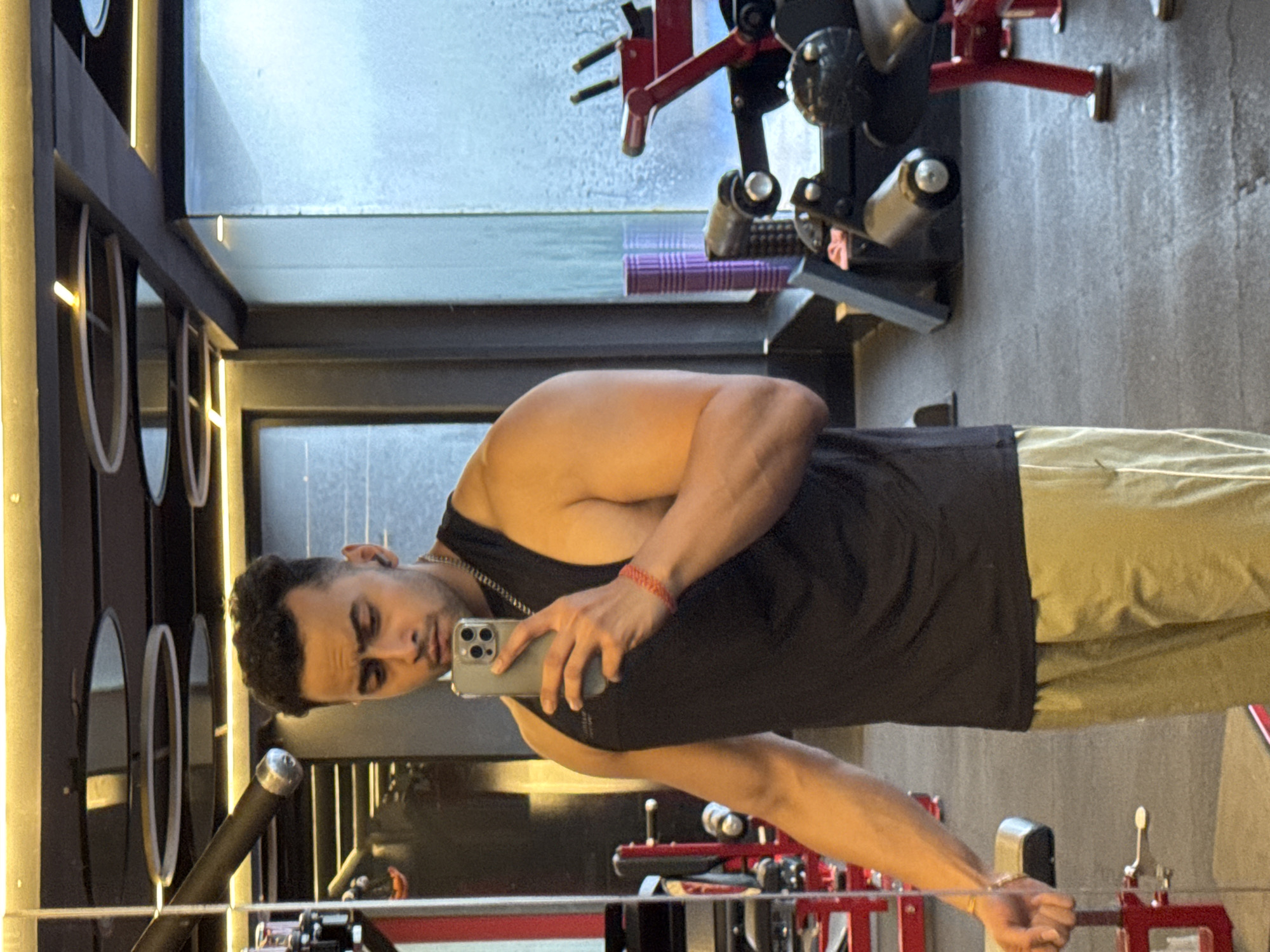 An athletic South Asian man with short, dark, curly hair and a light beard takes a mirror selfie in a gym. He is wearing a black sleeveless tank top and light brown athletic shorts. A silver chain is visible around his neck, and a red woven bracelet is on his right wrist. He holds a silver smartphone in his right hand, capturing his reflection. The gym environment is characterized by a dark ceiling with circular light fixtures and yellow strip lighting. Red and black weight training equipment, including a leg press machine, are visible in the background, along with a frosted glass window. The man appears to be standing in a gym, possibly in Kolkata, India, given the context.