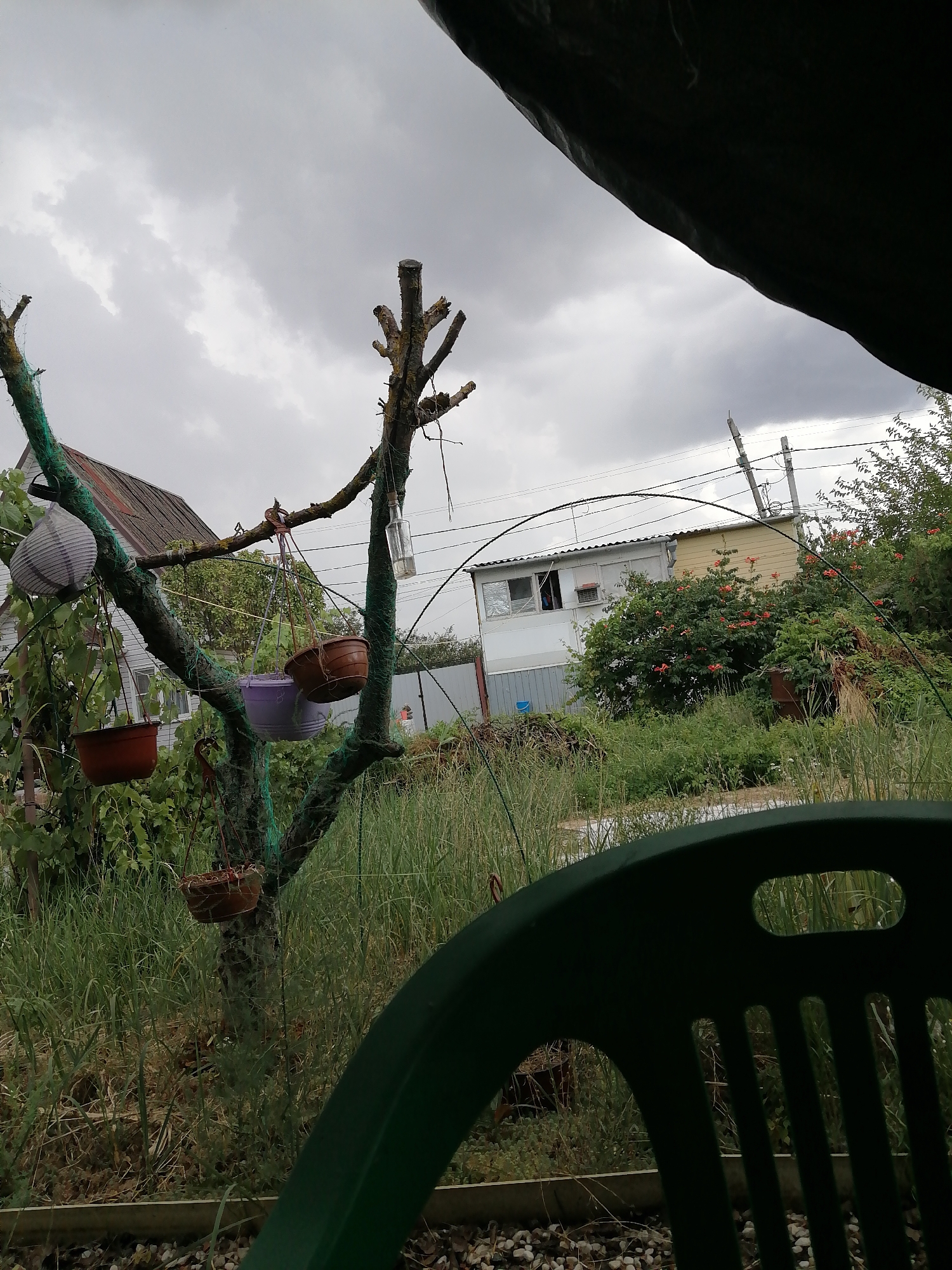 The image depicts an outdoor scene, likely a backyard or garden area. In the foreground, a dark green plastic chair is partially visible, obscuring the view of the ground which is covered with gravel. Beyond the chair, a tree with several branches extends upwards, adorned with hanging flower pots of various colors (red, purple, and brown) and a decorative paper lantern. Some of the branches are wrapped with green netting. A house with a gray siding and a reddish roof is visible in the background, along with another smaller, light-colored building. Lush green grass and foliage surround the area. Power lines and utility poles are present in the distance, suggesting an urban or suburban setting. The sky is overcast with heavy gray clouds. There are no identifiable people present in the scene. The location is identified as Rostov-on-Don, Russia, indicating a potentially dacha or country house setting.