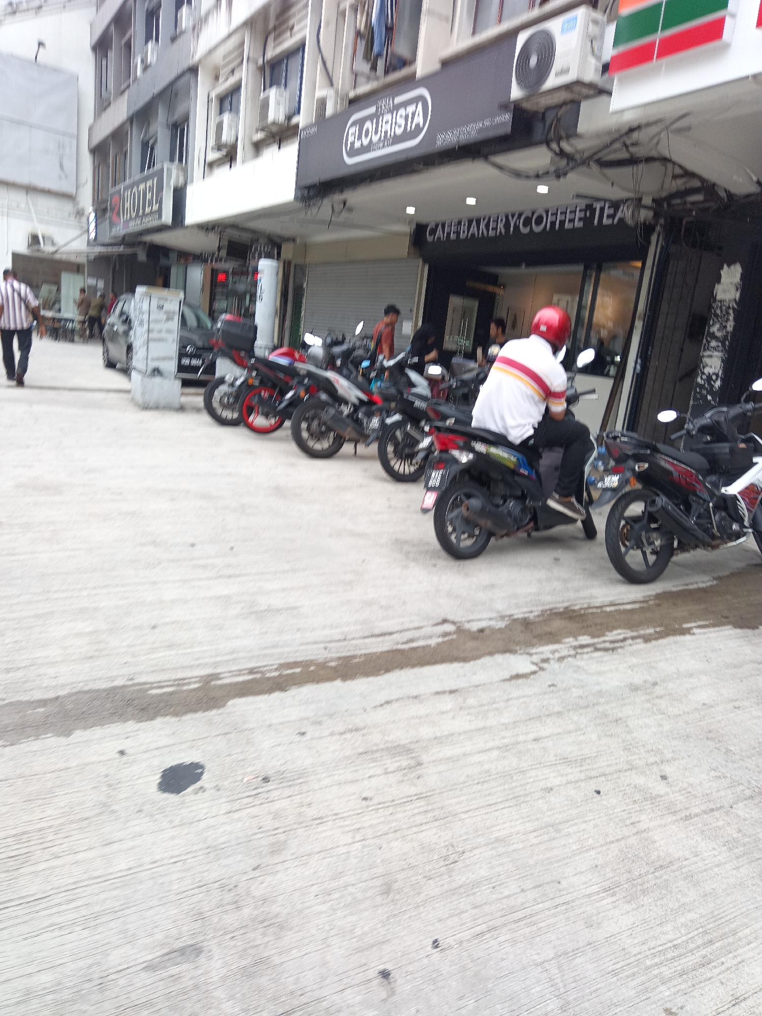 The image depicts a street scene in Kuala Lumpur, Malaysia, specifically in front of a commercial building. A person is walking on the left side of the frame, wearing a striped shirt and dark pants. Several parked motorcycles are visible, with one rider wearing a red helmet and a white and red striped shirt, sitting on a motorcycle. The building behind the motorcycles features signage, including "2 HOTEL," "FLOURISTA," and "CAFE BAKERY COFFEE TEA." The street is paved with concrete, and there are some wet patches on the ground. The overall scene suggests a commercial area with shops and services.