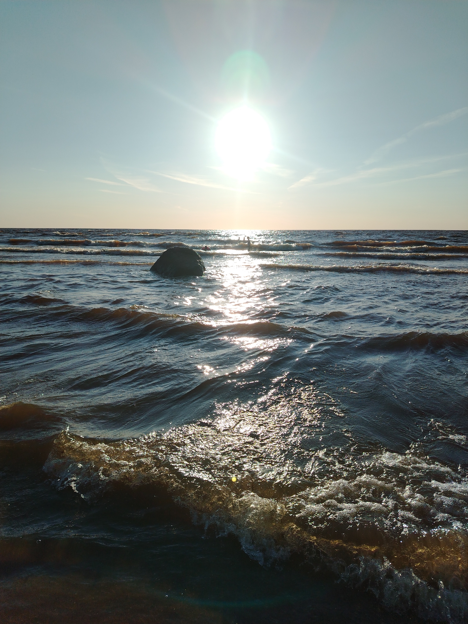 The image depicts a sun-drenched ocean scene with gentle waves rolling towards the shore. The sun, positioned high in the clear sky, casts a brilliant glare on the water, creating a shimmering effect. A large, dark rock stands partially submerged in the sea, adding a natural element to the landscape. The horizon line is distinct, separating the water from the sky, which is a pale blue with a few wispy clouds. The overall atmosphere is tranquil and serene, evoking a sense of peace and natural beauty. The location of Ivanovskoe, Russia, is not depicted in the image.