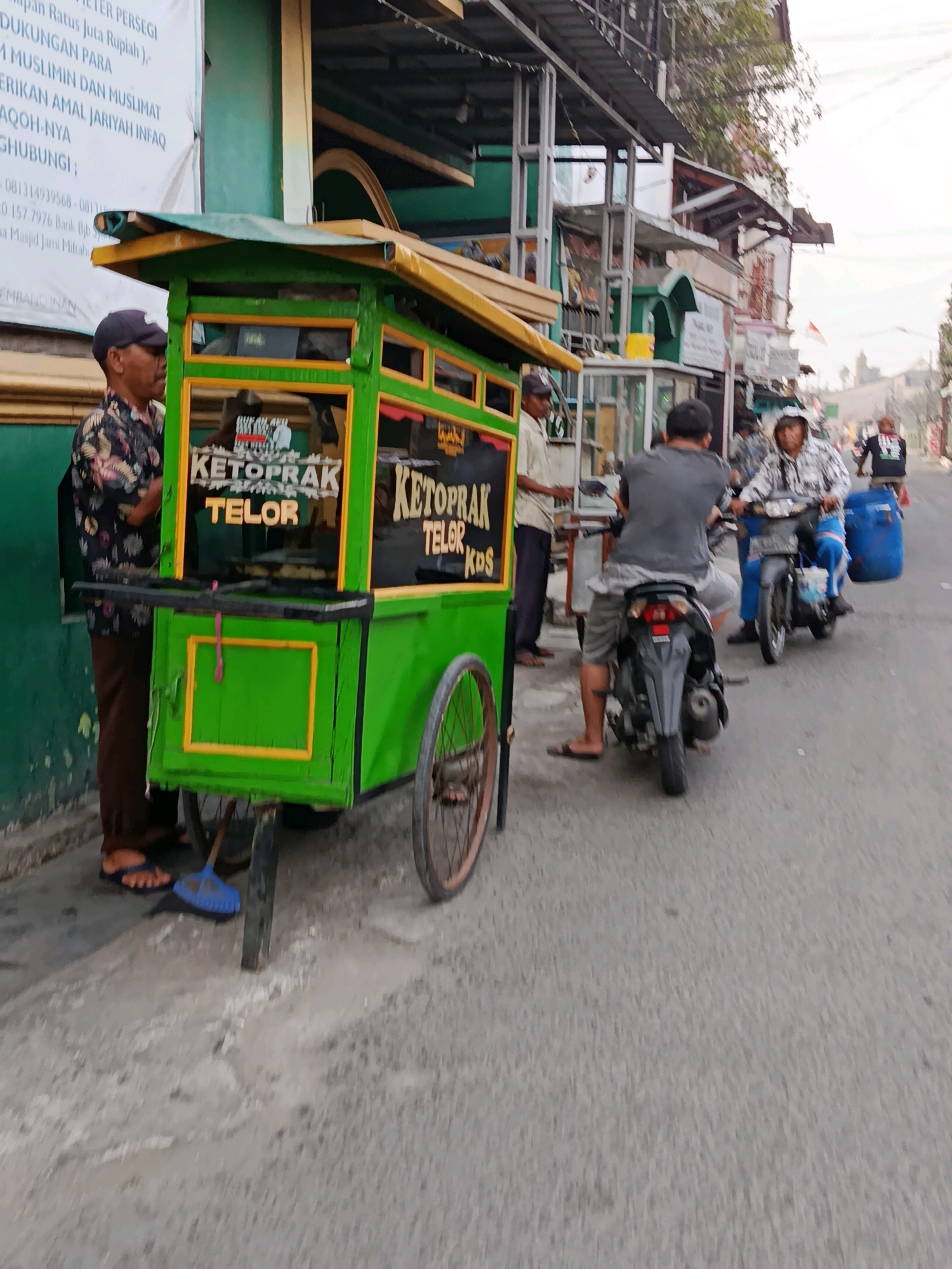 The image depicts a street scene in Bekasi, Indonesia, likely in an urban area with commercial establishments. In the foreground, a vibrant green food cart is prominently displayed, advertising "KETOPRAK TELOR KDS". A man wearing a patterned shirt and a dark cap stands next to the cart, appearing to be an attendant or owner. To the right of the food cart, two men are seated on motorcycles. The first man, facing away from the camera, is wearing a grey shirt and shorts. The second man, also on a motorcycle, is wearing a patterned jacket and a beanie, with a large blue barrel behind him. Further in the background, other individuals and motorcycles are visible, suggesting a bustling street environment. Buildings with signs and awnings line the street, indicating shops or residences. The overall lighting suggests it is daytime, with possibly overcast conditions.