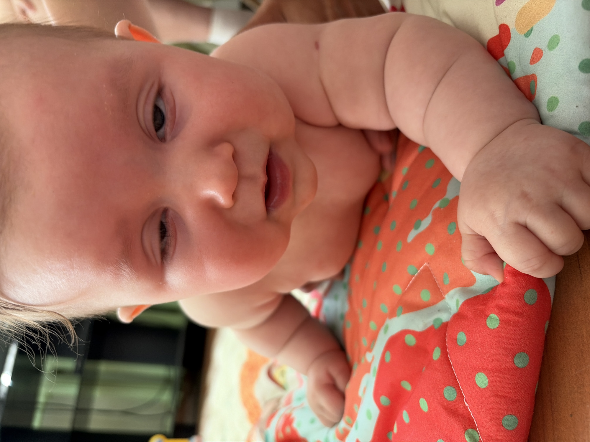 A baby is lying on its stomach on a colorful quilted blanket with red and light blue patches, each adorned with green polka dots. The baby's skin is fair, and the baby's mouth is slightly open. The image is taken at a close-up, high-angle view, and appears to be taken at an indoor location. There is no indication of Chaikovsky, Russia, in this image.