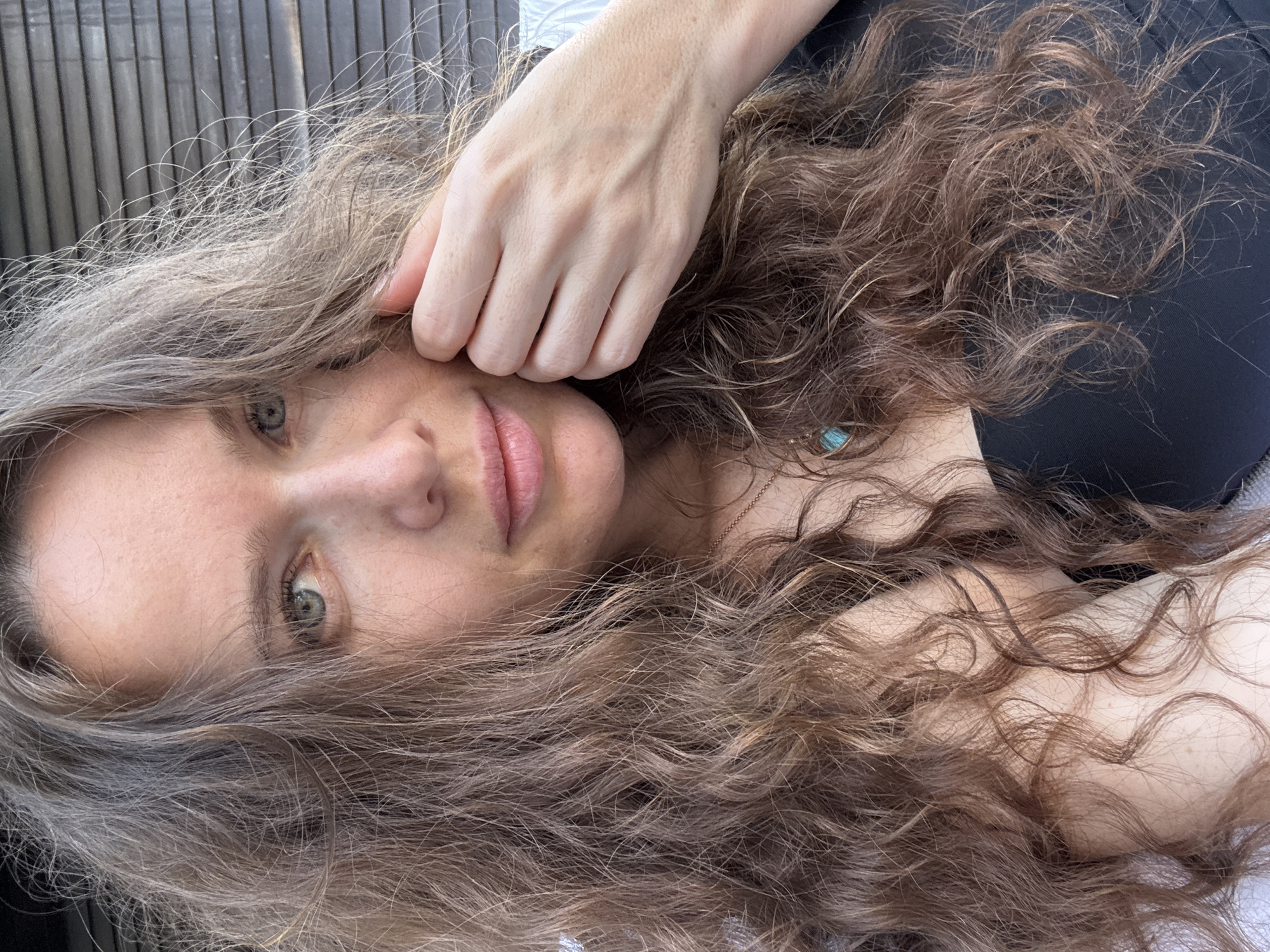 A medium close-up selfie shows a woman with long, wavy brown hair. She is looking towards the camera with her right hand near her face. She is wearing a black top and a delicate necklace with a turquoise pendant. The background consists of horizontal wooden slats. The lighting suggests it is daytime. The woman appears to be outdoors, possibly on a balcony or patio. The specific location in Dubai is not discernible from the image alone.
